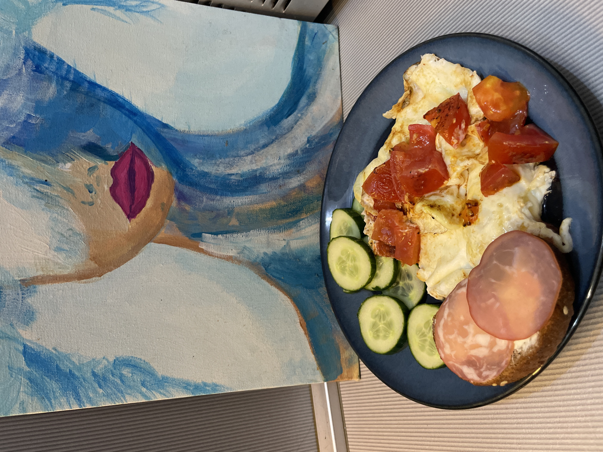 An abstract painting of a woman's face with blue hair and bright pink lips is displayed behind a dark blue plate of food. The plate contains an omelet topped with chopped tomatoes, slices of cucumber, and an open-faced sandwich with cream cheese and thinly sliced salami. The painting is on a white canvas with visible brushstrokes. The background is a ribbed surface in shades of gray and white. No information about the location in Sochi, Russia, is provided in the image.