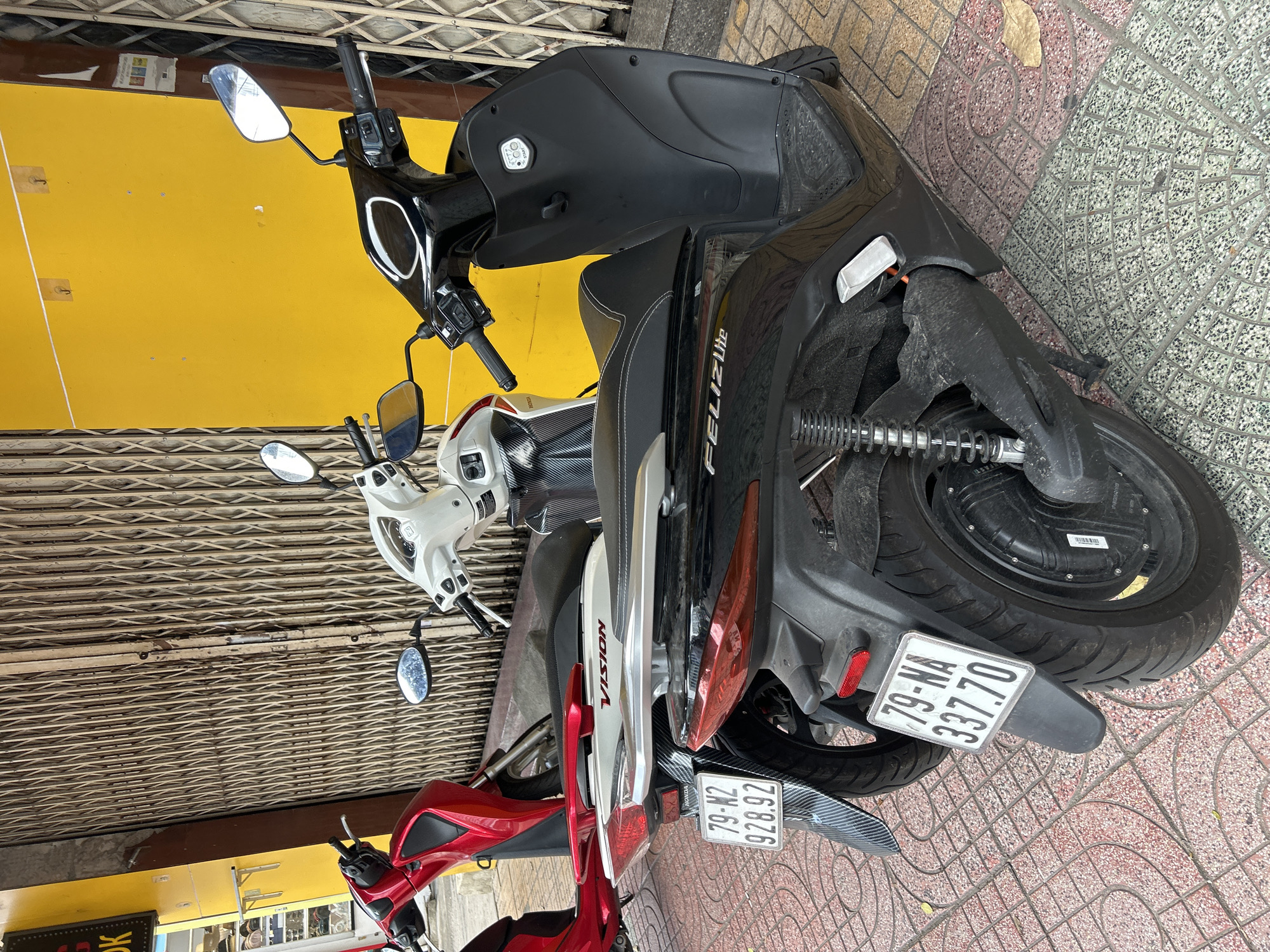Three scooters are parked on a tiled sidewalk next to a building with a bright yellow wall and a closed metal rolling shutter. The image is taken during the daytime.
The most prominent scooter, positioned on the right, is black and appears to be an electric model, indicated by the hub motor visible on the rear wheel. The text "FELIZ Lite" is visible on its side. Its rear license plate reads "79-NA 337.70".
Behind the black scooter and slightly to the left, a white scooter is parked. The word "VISION" is visible on its side, identifying it as a Honda Vision model.
Further to the left and partially obscured by the other scooters, a red scooter is visible. Its rear license plate, "79-N2 928.92", is clearly readable. A small "HONDA" logo is also visible near this license plate.
The sidewalk features patterned red and grey tiles. A single dried leaf rests on the tiled ground. The yellow wall behind the scooters has some faint white markings and a small, unidentifiable paper attached to it. Above the yellow wall is a corrugated metal rolling shutter, common for storefronts. The license plates, with the '79' provincial code, indicate the setting is Khánh Hòa province, Vietnam, consistent with the given location context of Nha Trang.