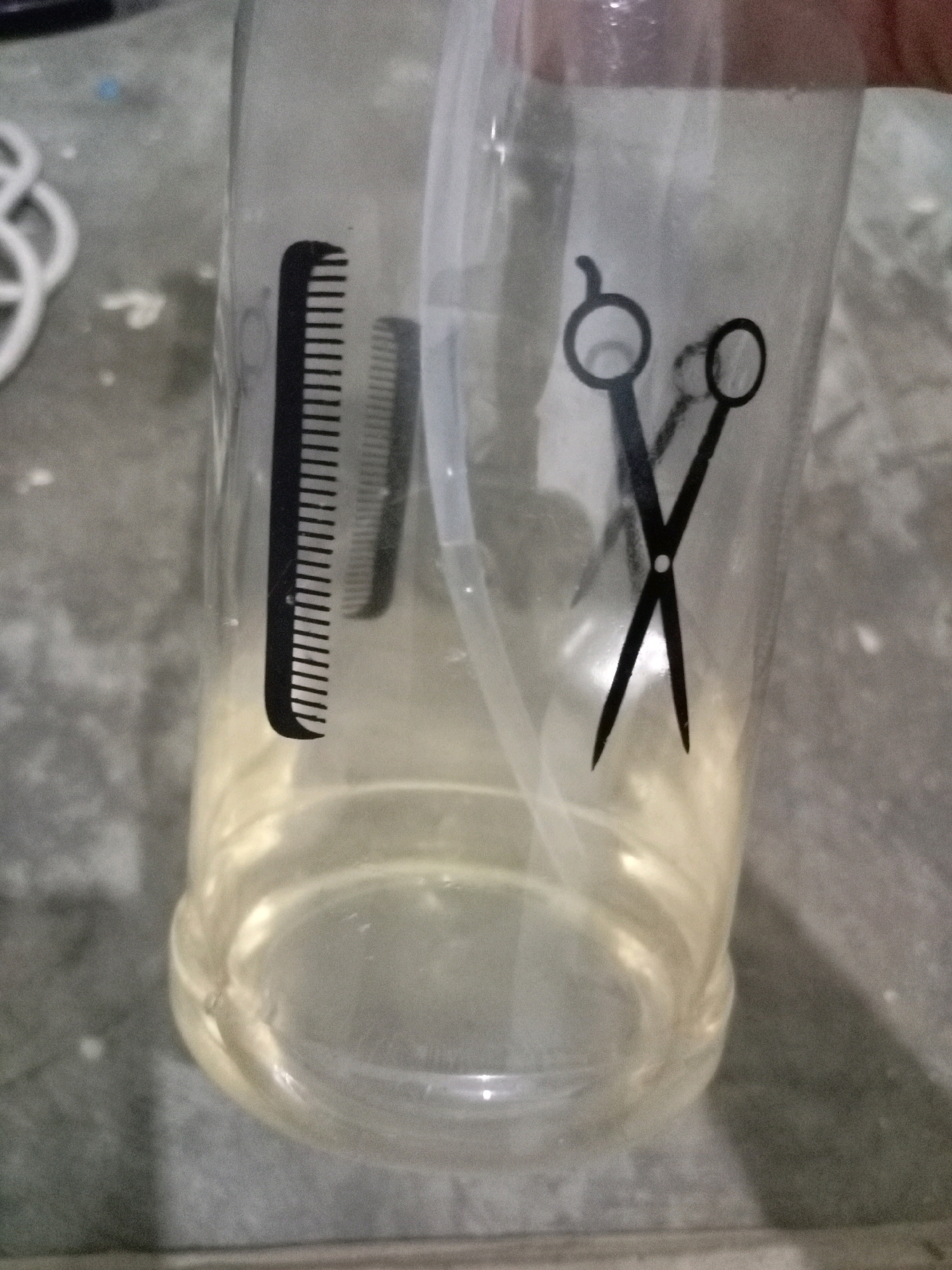 The image shows a clear plastic spray bottle containing a yellow liquid. The bottle has black decals of a comb and scissors on its exterior. A white tube extends from the neck of the bottle into the liquid. The bottle is placed on a gray, concrete-like surface. The background is blurred and appears to be indoors, possibly a workshop or salon. There are no people visible in the image. The lighting suggests an indoor environment. The image does not provide any specific information about its location in Batam, Indonesia.