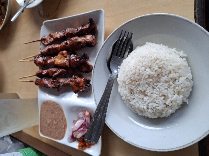 A white plate holds a circular mound of fluffy white rice, with a fork and spoon resting on top. Beside it, another white plate features four satay skewers with grilled meat, accompanied by a small portion of peanut sauce and a side of sliced red onions with chili. The overall presentation suggests a meal, likely Asian cuisine.