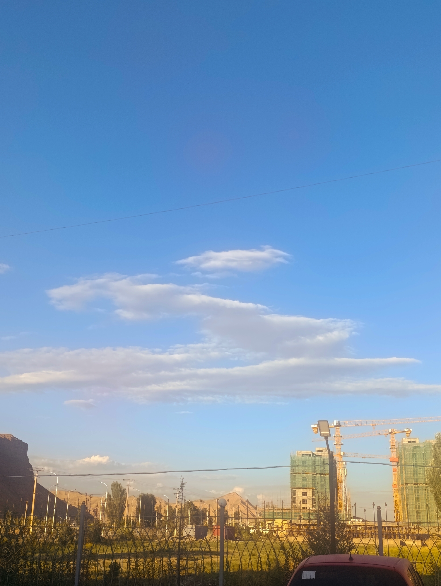 This image captures a scene in the city of Naryn, Kyrgyzstan. The sky is a clear, bright blue with scattered white clouds. In the mid-ground, two large construction cranes are visible, one yellow and one orange, towering over buildings under construction that are wrapped in green netting. A streetlamp stands in the foreground. A metal fence runs across the lower part of the image, separating the viewer from a grassy area with some trees and more distant, possibly unfinished, buildings. A large, dark red vehicle is partially visible in the bottom right corner. The background features a large, steep, rocky hill on the left side. The overall impression is one of ongoing development within a semi-arid or mountainous landscape.