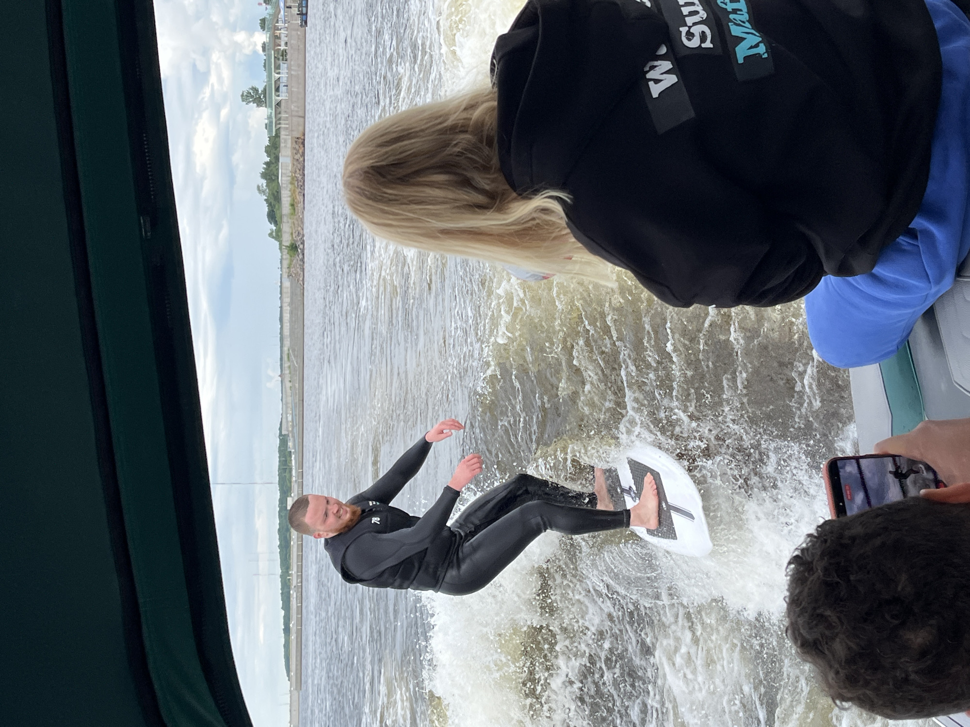 A man in a black wetsuit is wake surfing on a white board behind a boat. He is leaning back, with his arms outstretched, and his eyes are closed. The water is choppy, and there is a lot of spray. In the background, a person with blonde hair is visible, and another person's hand is holding a phone, presumably to take a picture. The sky is cloudy, suggesting it might be a partly cloudy day. The scene appears to be a recreational activity on a body of water, possibly a lake or a river, given the context of Saint Petersburg, Russia. The overall atmosphere seems active and adventurous. The visible text on the person's jacket reads "W. Surf."