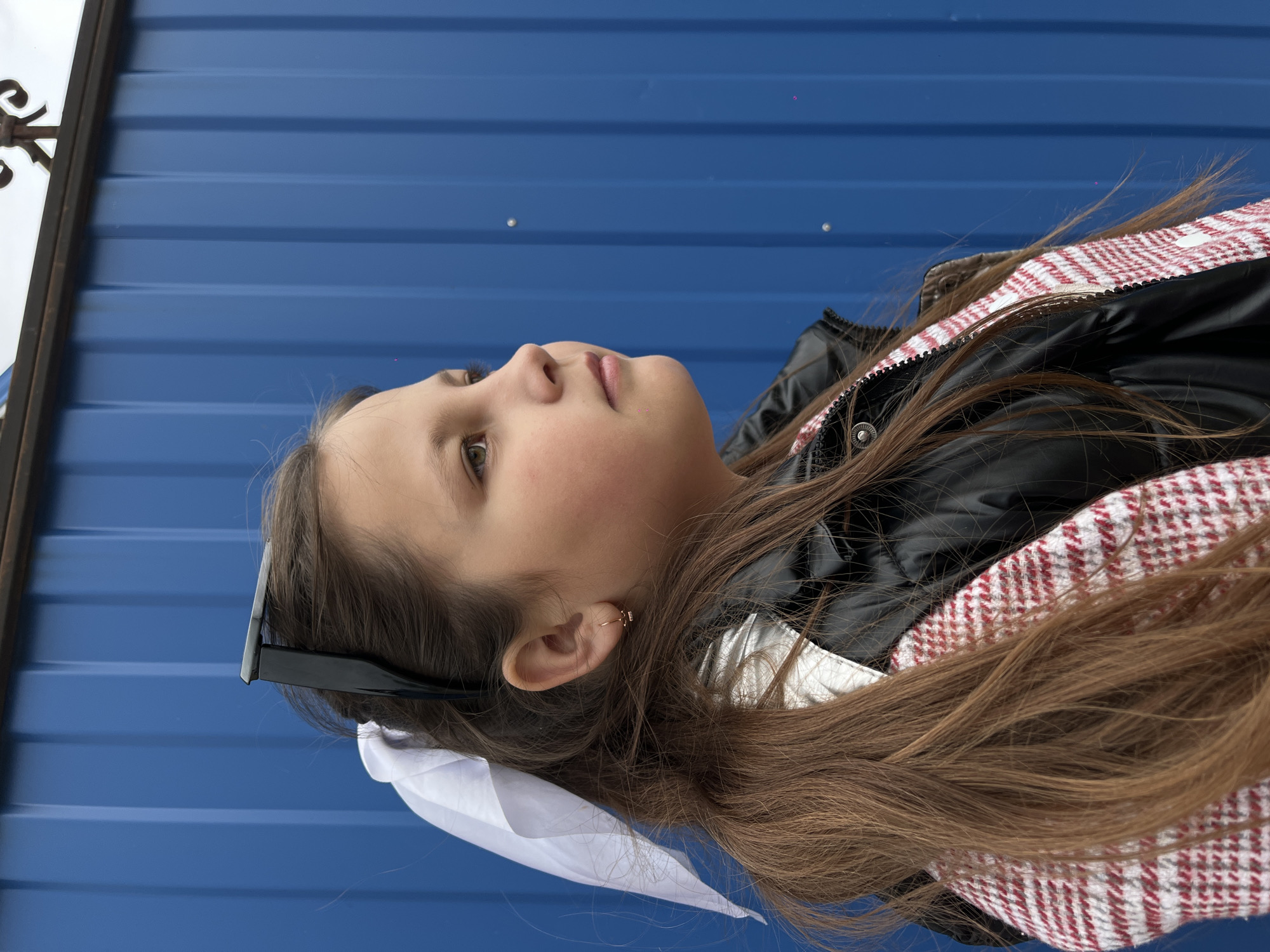 A portrait-oriented close-up features a young girl, possibly a pre-teen, looking upwards with a contemplative expression. Her head is angled towards the bottom left of the frame, with her gaze directed towards the upper right. She has long, straight brown hair that cascades over her shoulders and down her back. A pair of black sunglasses rests on top of her head, pushed back into her hair. In her visible right ear, she wears a small, delicate gold hoop earring with a tiny embellishment.
She is dressed in a dark, likely black, puffer jacket. A distinctive red and white checkered or plaid pattern is visible on the inner lining or a decorative part of the jacket around her shoulder and collar area. Underneath her hair on the left side, a piece of white fabric, possibly a bow or part of her clothing, is visible.
The background consists of a solid, deep blue corrugated metal surface, likely a fence or wall, with horizontal ridges. The lighting suggests it is daytime, with soft, even illumination on her face. On the far left edge of the image, a small portion of an ornate, dark metal scrollwork design, possibly part of a gate or railing, is visible against a lighter background. There is no visible text in the image. The scene is set in Novopokrovskaya, Russia.