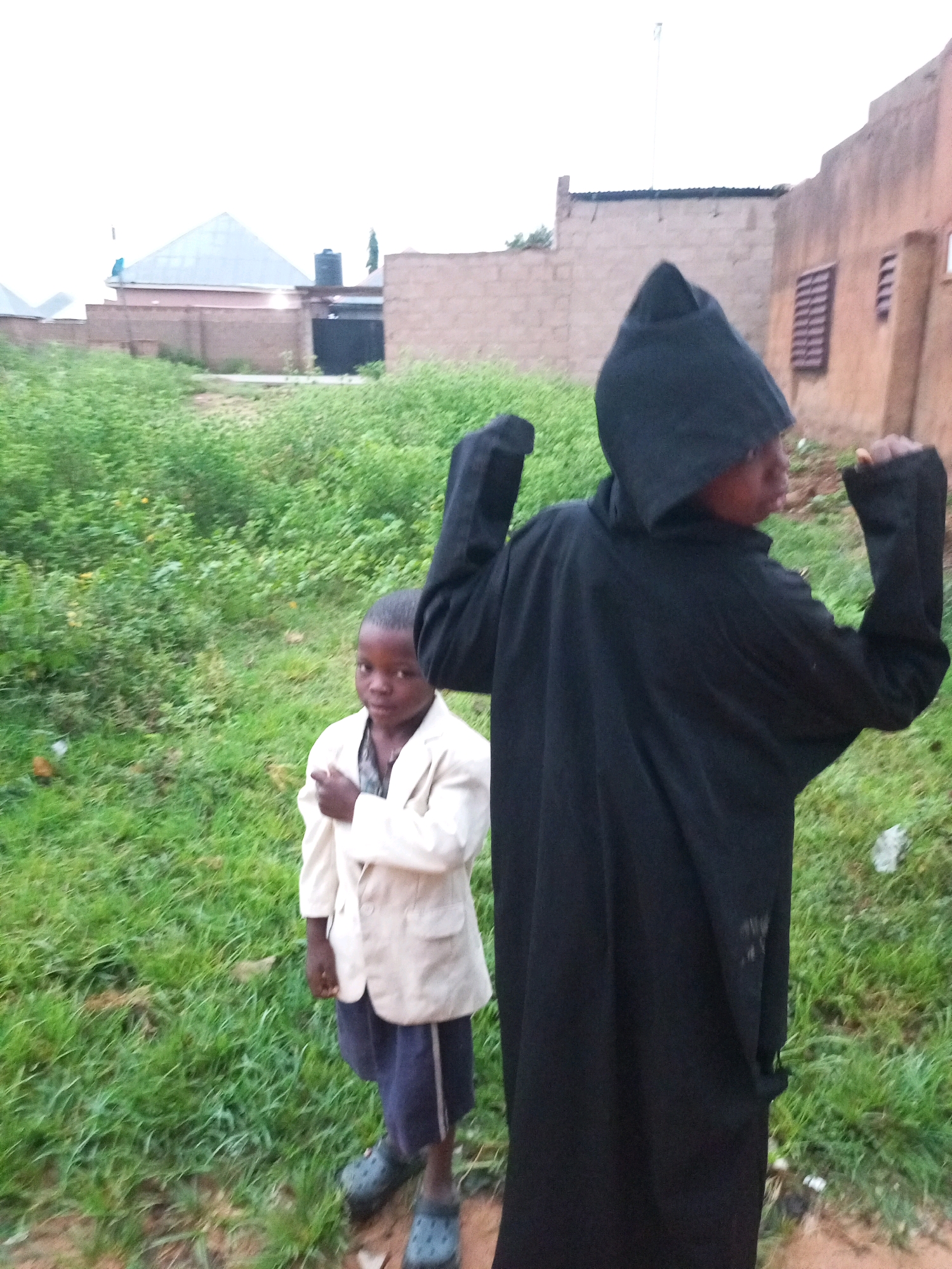 The image shows two children outdoors, possibly in the city of Tempure, Nigeria. On the left, a young boy is wearing a light-colored blazer over a darker shirt and navy shorts, with dark clogs on his feet. He is looking towards the camera with his right arm bent at the elbow, hand pointing slightly forward. To the right, a taller child, whose face is mostly obscured, is wearing a black hooded garment. This child has their arms raised and bent at the elbows, with fists clenched. They are facing away from the camera, looking to their right. The background consists of overgrown green grass and vegetation, with a brown building with small windows visible to the right and a partially visible building with a light-colored roof in the upper left. The ground appears to be dirt.