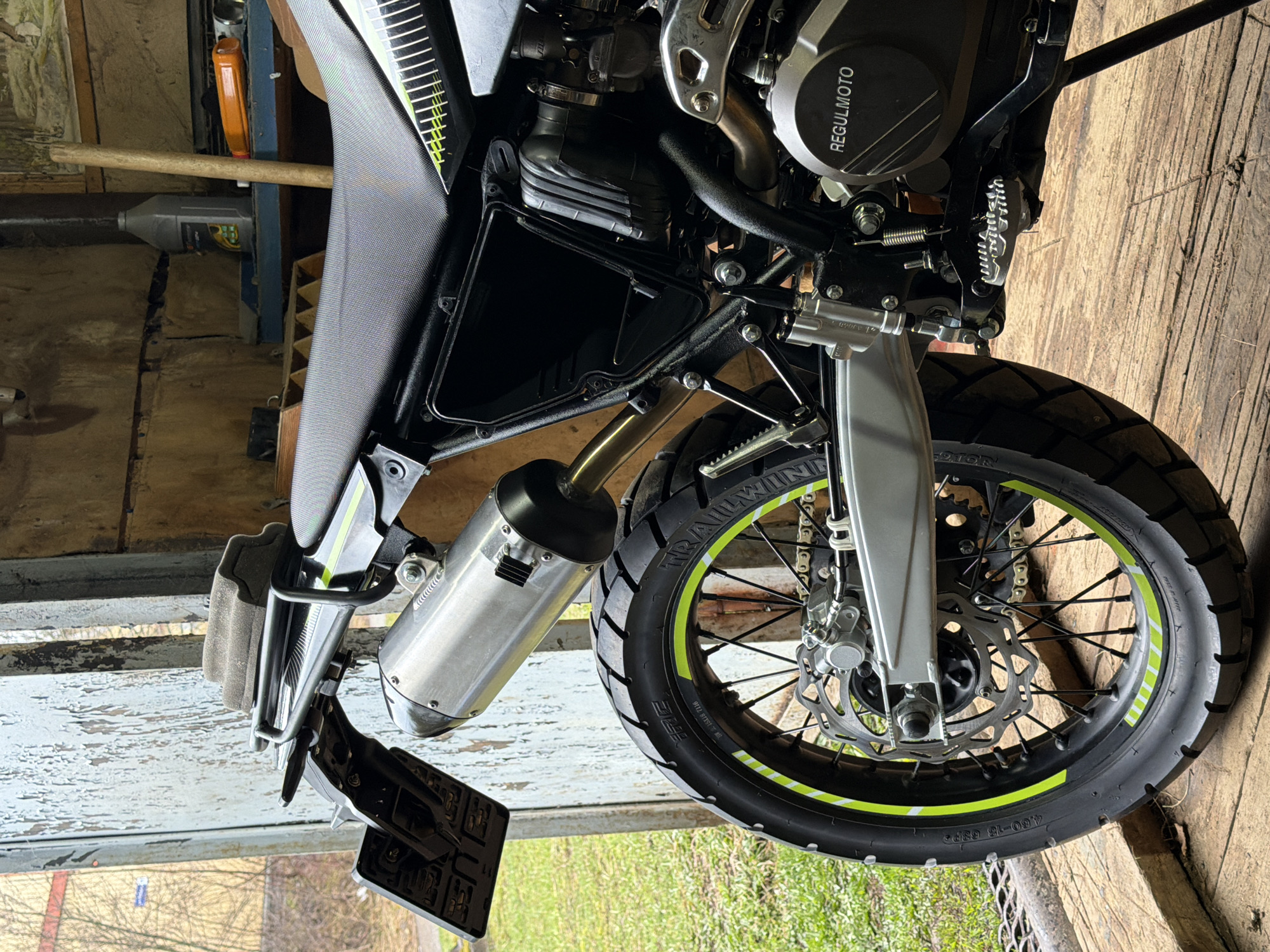 The image shows a close-up of a Regulmoto motorcycle. The motorcycle is predominantly black with lime green accents on the rims. Visible components include the front tire, front suspension, exhaust pipe, engine block, seat, and license plate holder. The background suggests an indoor space with a worn wooden floor and a partially visible window showing trees outside. Based on the motorcycle’s branding, the image is possibly taken in or related to Большой Букор, Russia. No people are visible.