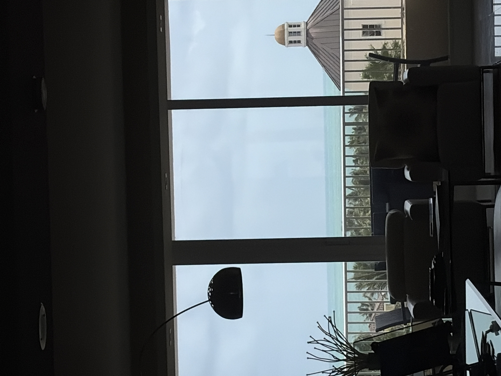 The image is taken from inside a building, looking out of a window towards what appears to be a beach and ocean view. A building with a dome on its roof is visible in the distance. The view outside the window includes palm trees and a railing. Indoors, there is a chair and a floor lamp visible. The location is identified as Key Biscayne, United States of America.