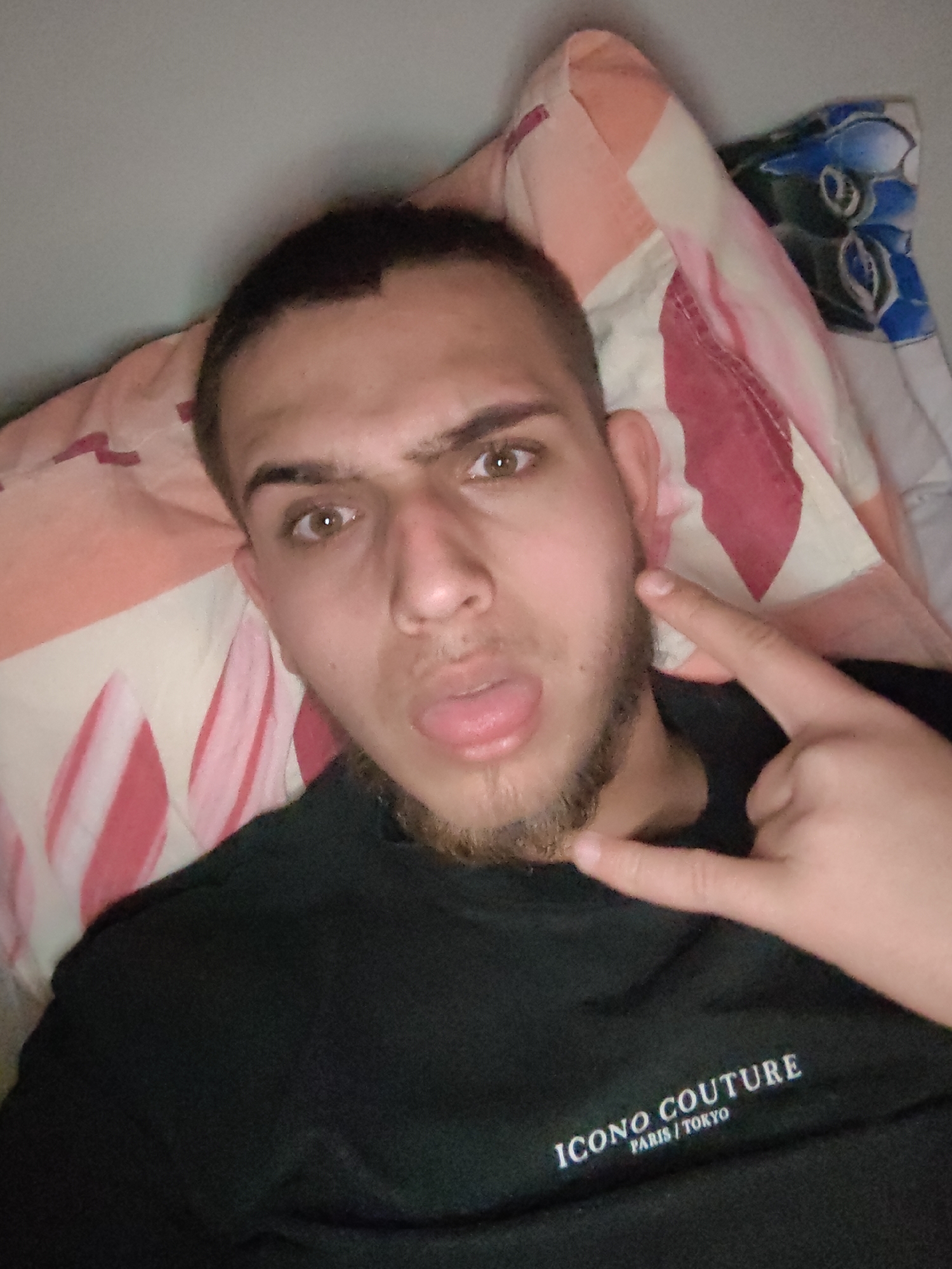 A young man with a short haircut and a beard is lying on a bed with patterned sheets. He is wearing a black t-shirt with the words "ICONO COUTURE PARIS / TOKYO" printed on it. He is sticking his tongue out and making a rock-and-roll gesture with his hand. The location is unknown, but the city is given as Sofia, Bulgaria.