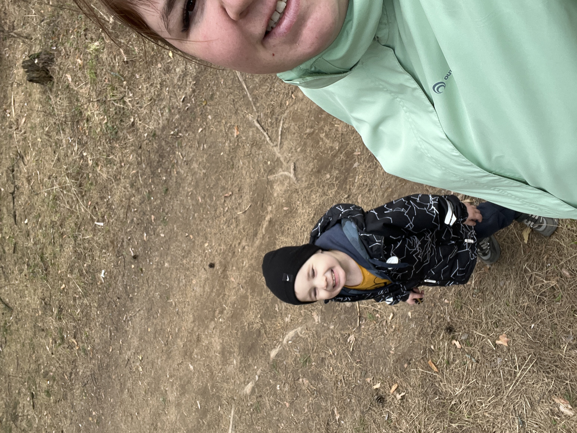 This overhead selfie-style photograph captures an adult and a young child outdoors, rotated on its side. The adult's face, smiling widely, occupies the top right portion of the frame, partially obscured. They are wearing a light mint-green jacket with a visible grey "OUT" logo and a circular design on the chest.
Below and to the left, the child stands, looking up at the camera with a bright, joyful smile. They are dressed in warm clothing suitable for cool weather: a black beanie featuring a white Nike swoosh, a yellow t-shirt, a blue underlayer, and a black jacket adorned with a white geometric pattern. Dark pants and sneakers complete their outfit.
The setting is an outdoor area, possibly a park or forest edge in Chaikovsky, Russia, characterized by ground covered in dry, brown grass, patches of dirt, and scattered leaves and small twigs. The natural light suggests it is daytime. The overall mood conveyed by the smiles is cheerful and happy, indicating a pleasant moment shared between the two individuals.