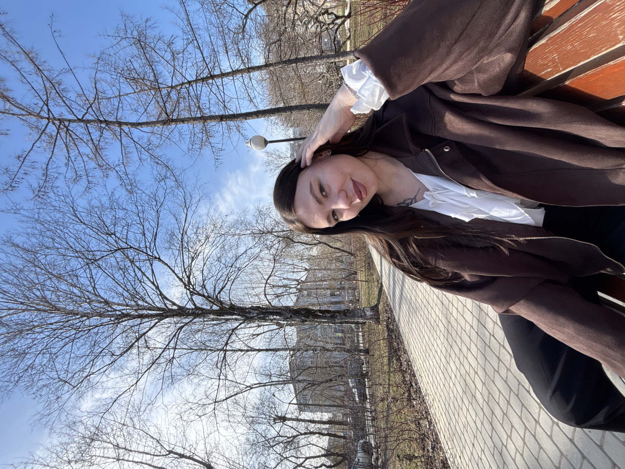 A young woman with long dark hair is seated on a park bench in what is likely the city of Perm, Russia. She is wearing a white blouse with lace detailing and a brown jacket. A portion of a tattoo is visible on her chest. The background features bare trees, a lamp post, and buildings typical of urban architecture found in Russia. The ground is paved with cobblestones. The sky is blue with some clouds.