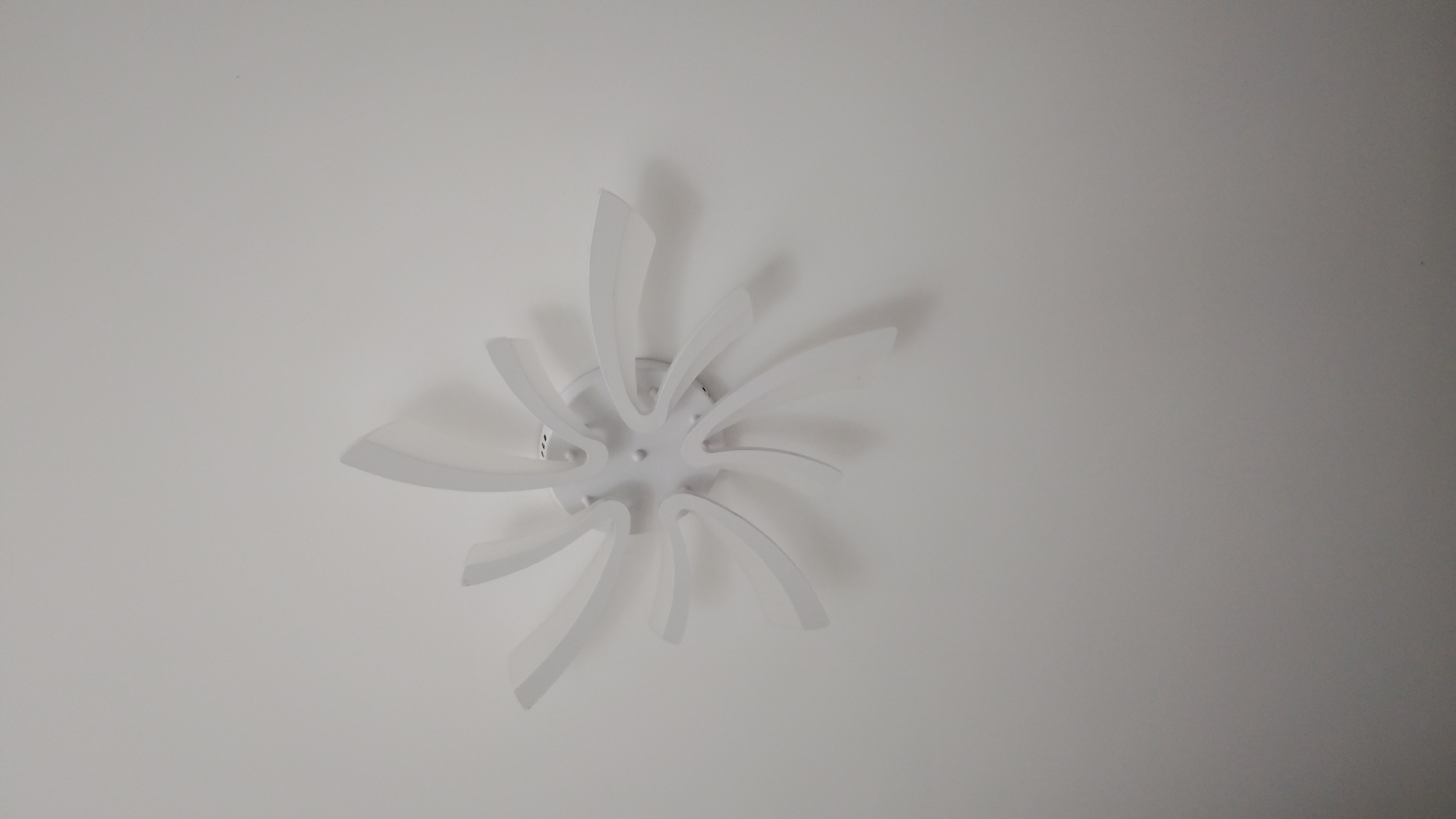 A modern, white ceiling light fixture is mounted on a white ceiling. The fixture has a central hub with several curved, petal-like elements radiating outwards, resembling a stylized flower or starburst. The lighting is uniform, suggesting it is either daytime with ambient light or the fixture itself is illuminated. There are no people or other objects visible in the image. The scene appears to be indoors, in a room with a white ceiling. There is no information available to specify the location within the city of Smolyan, Bulgaria.
