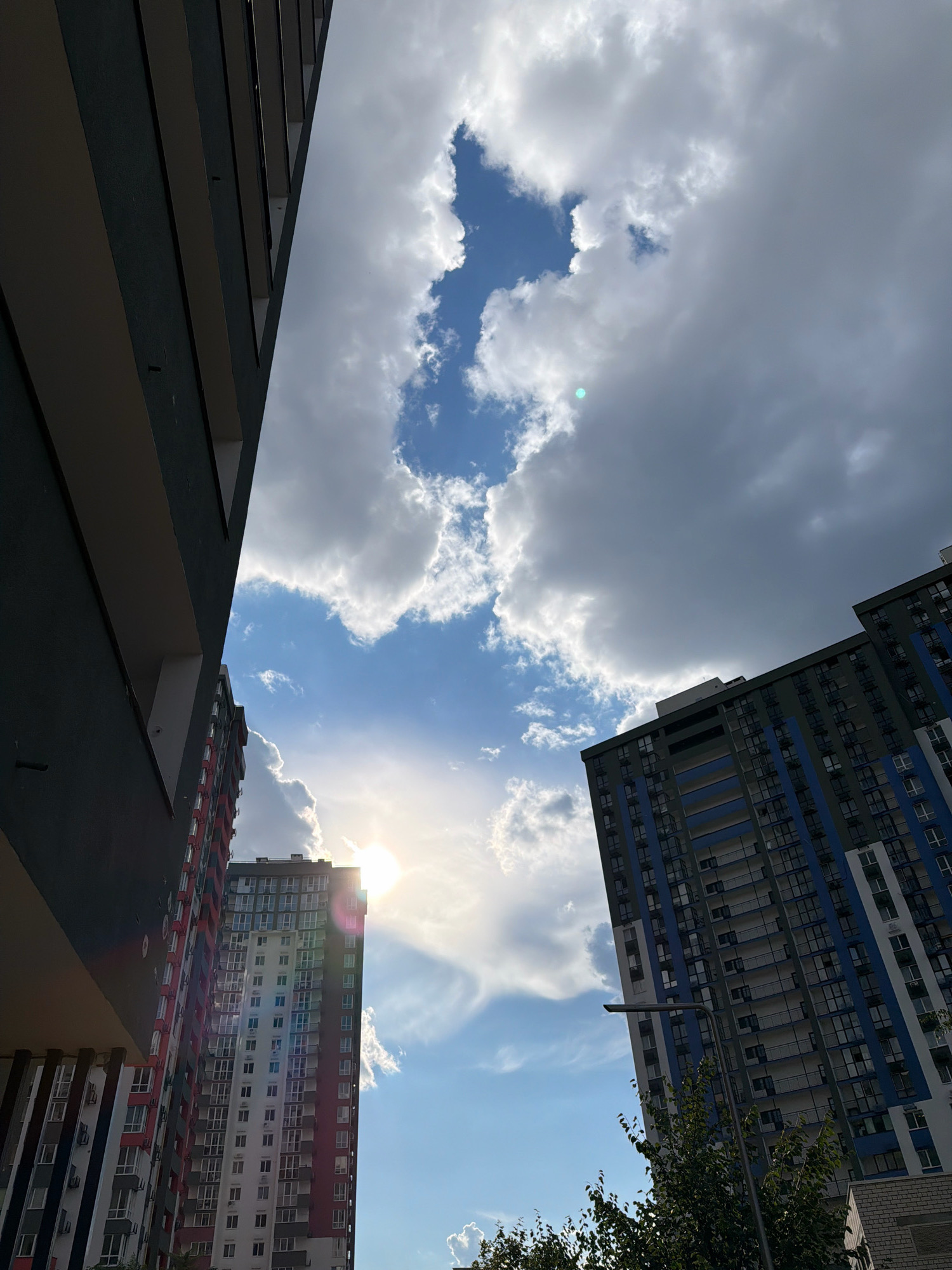 The image captures a low-angle view of modern high-rise apartment buildings against a cloudy sky. The buildings are characterized by their multi-story structures, featuring numerous windows and balconies. One building on the left is predominantly dark gray with lighter horizontal accents, while another on the right is tall and slender with a facade of blue and white. Sunlight breaks through a gap in the clouds, creating a bright flare and casting a lens flare effect on the left side of the frame. The sky is a mix of bright blue patches and large, grey clouds, indicating dynamic weather conditions. Trees and a lamppost are visible in the foreground, suggesting an urban residential area. The overall impression is one of contemporary urban architecture under a dramatic sky.