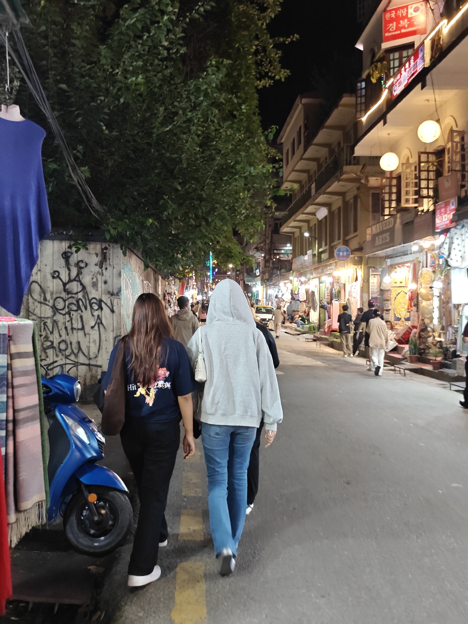 A night scene on a street in Kathmandu Metropolitan City, Nepal shows a paved road lined with multi-story buildings housing shops. A blue scooter is parked on the left side of the road, next to a wall covered in graffiti. Two women are walking away from the camera down the street. The woman on the left has long brown hair and is wearing a dark blue t-shirt and black pants. The woman on the right is wearing a gray hooded sweatshirt and blue jeans. Several other people are walking further down the street, and shops on the right side of the road are illuminated with lights and display various goods. A sign with Korean characters and a Korean flag is visible on one of the buildings, indicating a Korean restaurant. Trees are visible on the left side of the street, and the sky is dark.