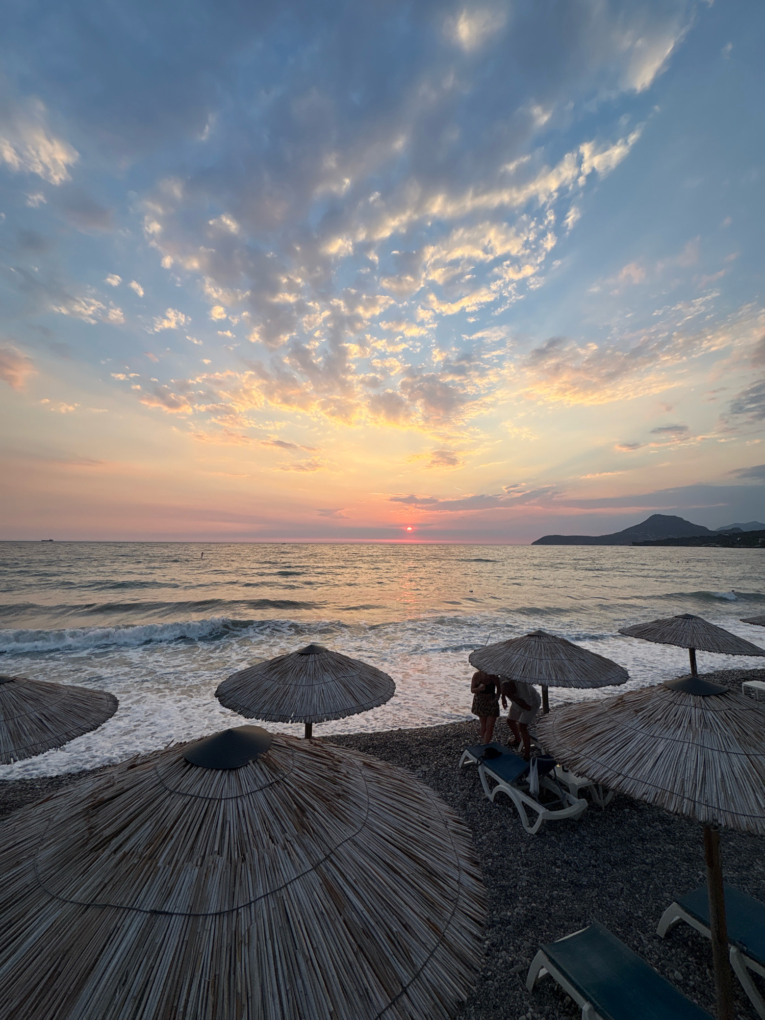 The image depicts a sunset on a pebbled beach in Bar, Montenegro. The sky is filled with a mix of blue and pink clouds, illuminated by the setting sun which is visible as a small red orb on the horizon over the Adriatic Sea. The sea is calm with gentle waves breaking on the shore, creating white foam. In the foreground, several thatched beach umbrellas provide shade over empty lounge chairs. Two people, one in a patterned dress and the other in a light-colored shirt and shorts, are standing near two lounge chairs under one of the umbrellas. Further out in the sea, a distant ship is visible on the left, and a lone figure can be seen on a paddleboard. The shoreline extends into the distance to the right, where a dark, mountainous landscape is silhouetted against the colorful sky. The overall scene conveys a peaceful and serene atmosphere of a beach at dusk.