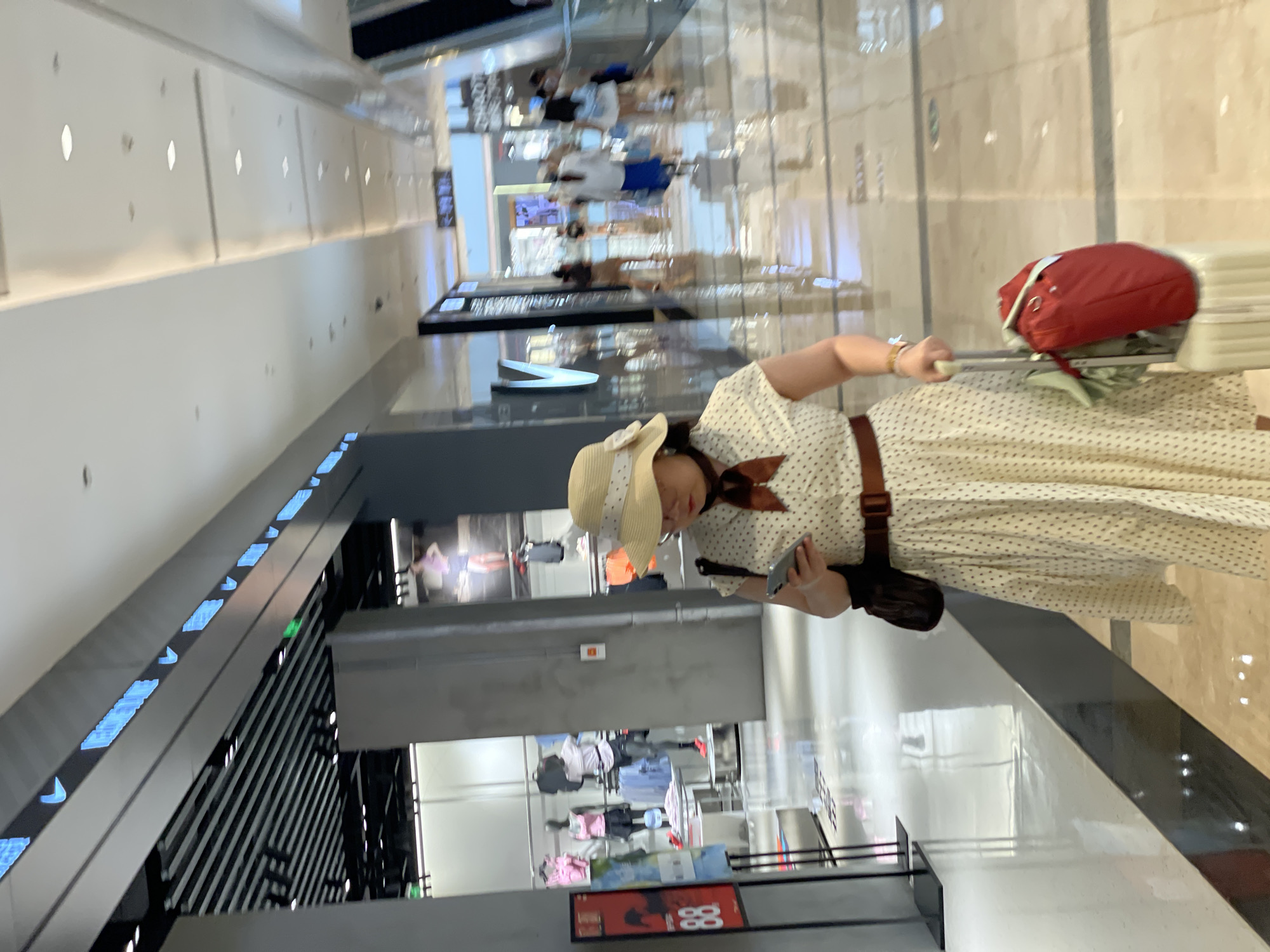In Yuzhong District, China, a woman in a light-colored, polka-dotted dress, a wide-brimmed straw hat with a white ribbon, and a brown scarf is walking through a modern shopping mall. She is holding a smartphone in her right hand and a light-colored wheeled suitcase with a red bag and a green garment attached to it with her left. The mall features a polished, reflective floor and a bright, contemporary interior with shops visible in the background. The woman is facing towards the right of the frame, looking down at her phone. The scene captures a moment of transit or leisure within an urban commercial space.