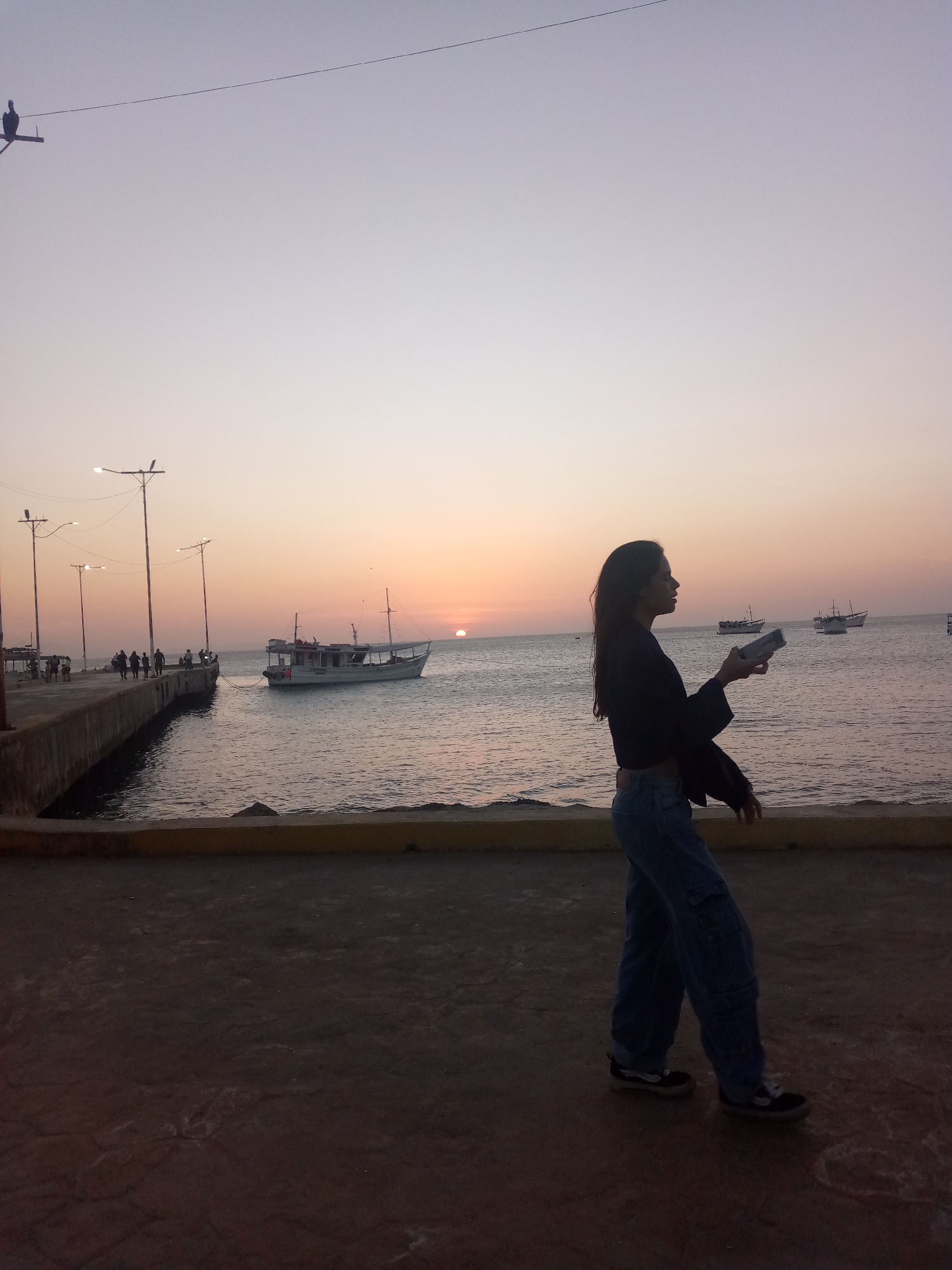 A young woman stands facing the sea at dusk in Juan Griego, Venezuela. She is holding a rectangular object, possibly a phone, in her hands. The sky is a pale gradient of colors, with the sun setting on the horizon. The sea is calm with several boats visible. A pier with a group of people and streetlights extends into the water on the left side. The platform where the woman stands is flat and paved, with a low yellow wall at the edge facing the sea.