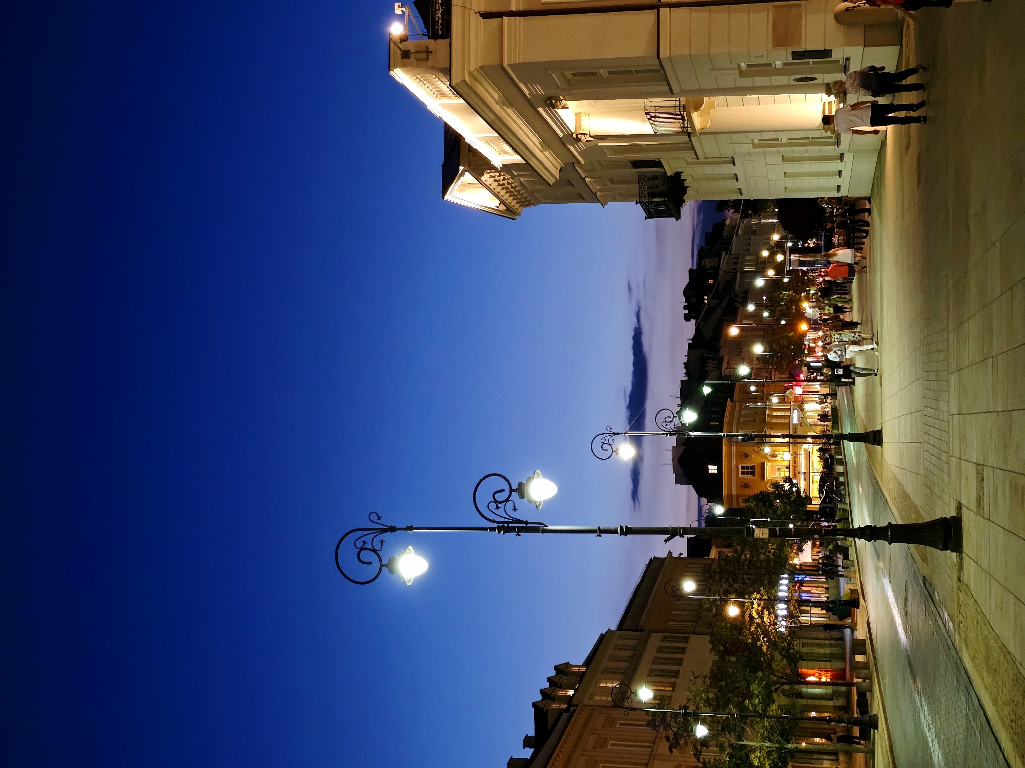 This image captures a street scene in Warsaw, Poland, during the twilight hours. The sky is a deep, clear blue, transitioning to hues of purple and pink near the horizon. Ornate, vintage-style streetlights with glowing bulbs illuminate the cobblestone street and the sidewalk.
On the right side of the street is a grand, neoclassical building with a white facade. It is lit from below, highlighting its architectural details like columns and a balcony with an iron railing. On the left side, a row of buildings with warm, lit windows suggests shops or restaurants. Trees with vibrant green foliage are interspersed along the sidewalk.
Several people are visible walking along the sidewalk. Some are strolling casually, while others appear to be in conversation. The street itself shows signs of light traffic, with a few cars present. The overall atmosphere is that of a lively urban evening.
