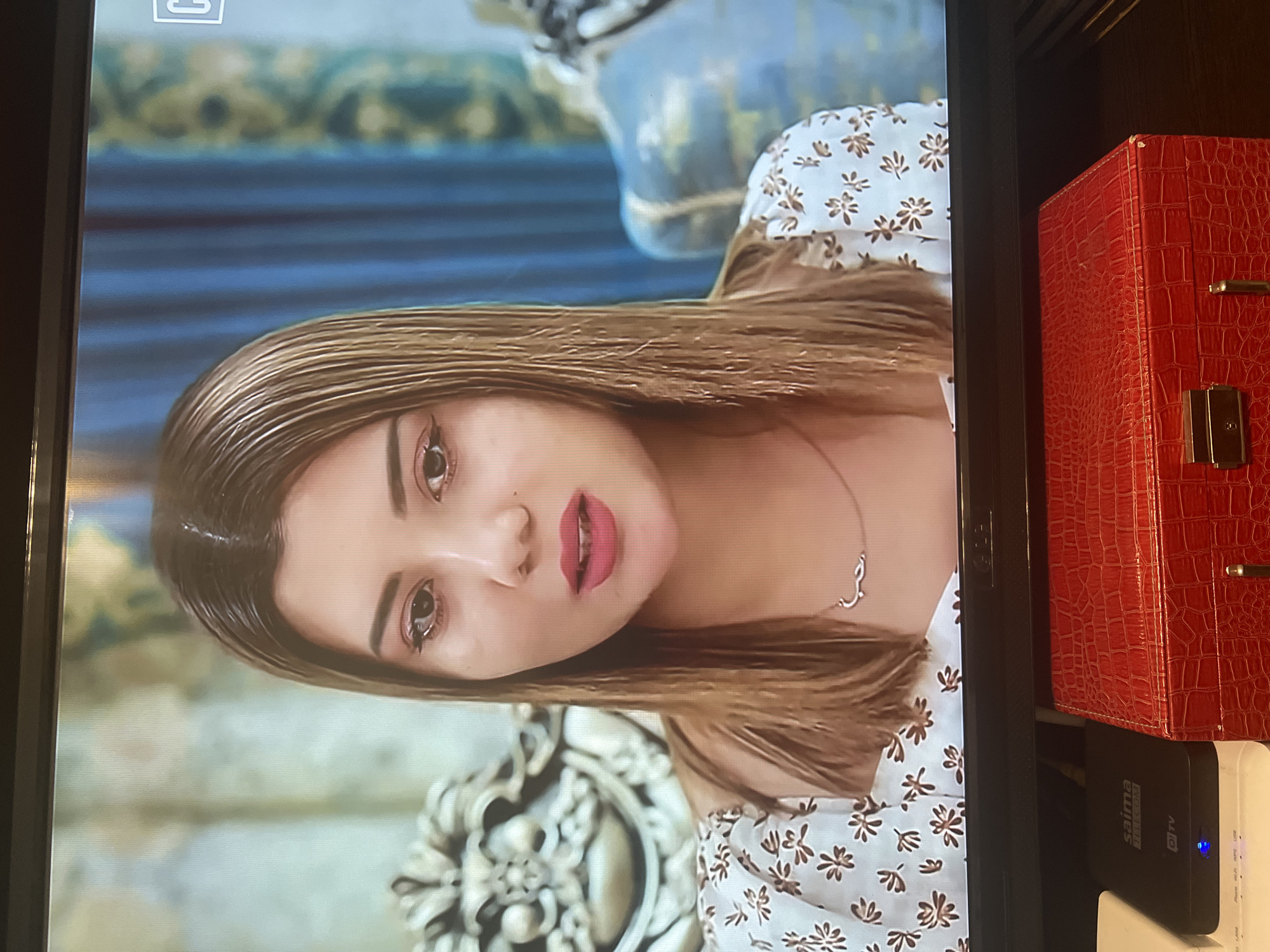 This is a medium close-up shot of a woman with shoulder-length, straight, light brown hair. She is wearing a white dress with a floral print of small brown flowers. She is also wearing a delicate gold necklace with an Arabic inscription. The woman's face is slightly turned to the side, and she appears to be speaking or looking at something off-camera. Her makeup is noticeable, with emphasis on her eyes and lips.
In the background, ornate furniture with carved details is visible. To the left, there's a light-colored, elaborate chair or sofa armrest. To the right, a blue curtain is partially visible, along with another piece of ornate furniture. The overall setting suggests an indoor environment, possibly a living room or studio.
Below the screen, there is a black electronic device with the "saima" logo and "OTTv" text. Adjacent to it is a red, textured box, possibly a jewelry box or storage case, with a metallic clasp. The image appears to be taken from a television screen, as indicated by the screen's frame and the presence of electronic devices below it.
The location is described as Osh City, Kyrgyzstan, but there are no specific visual cues in the image that directly confirm this location. The visual elements are generic to many indoor settings.
