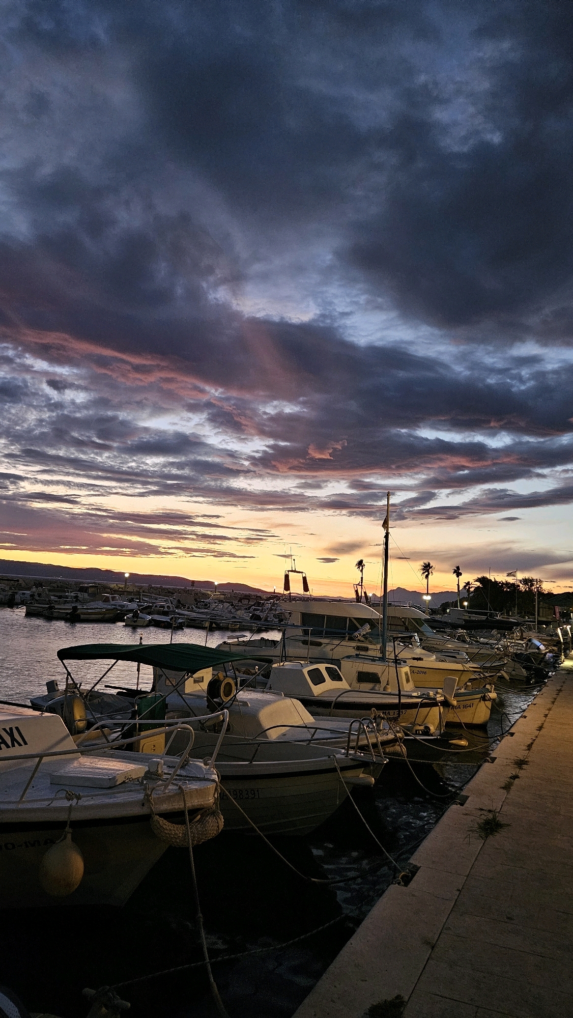 This image depicts a serene harbor scene at dusk in Općina Tučepi, Croatia. The sky is a dramatic canvas of deep blues and grays, streaked with vibrant pink and orange hues from the setting sun. Wispy clouds are illuminated by the last rays of light, creating a painterly effect.
The foreground is dominated by a row of docked boats, their hulls varying in shades of white and light yellow. Some boats have green canvas covers, while others are open. Ropes tether them to the dock, and a few buoys are visible, one bright yellow and round. The water is calm, reflecting the subdued colors of the sky.
To the right of the boats, a concrete pathway runs along the harbor. The pathway is illuminated by warm, artificial lights, casting a soft glow on the stone surface. Patches of green grass sprout from the cracks in the pathway, adding a touch of nature to the urban setting.
In the distance, a line of hills or mountains forms a dark silhouette against the horizon. The faint lights of buildings are visible along the coastline. The overall impression is one of tranquility and the peaceful closure of a day.