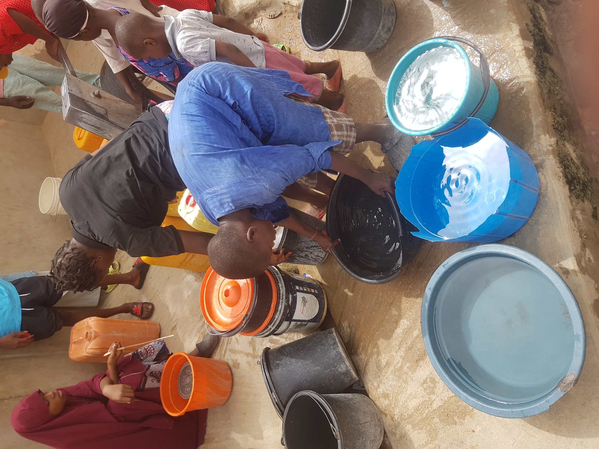 The image shows a group of people in Dutsinma A, Nigeria, collecting water. Several individuals are positioned around a water source, filling various buckets and containers. A child in a blue outfit is actively filling a black bucket. Other containers nearby include black, blue, and orange buckets. Some are filled with water. One woman is sitting beside a container. The ground around the water source appears wet, indicating recent spillage or ongoing activity.