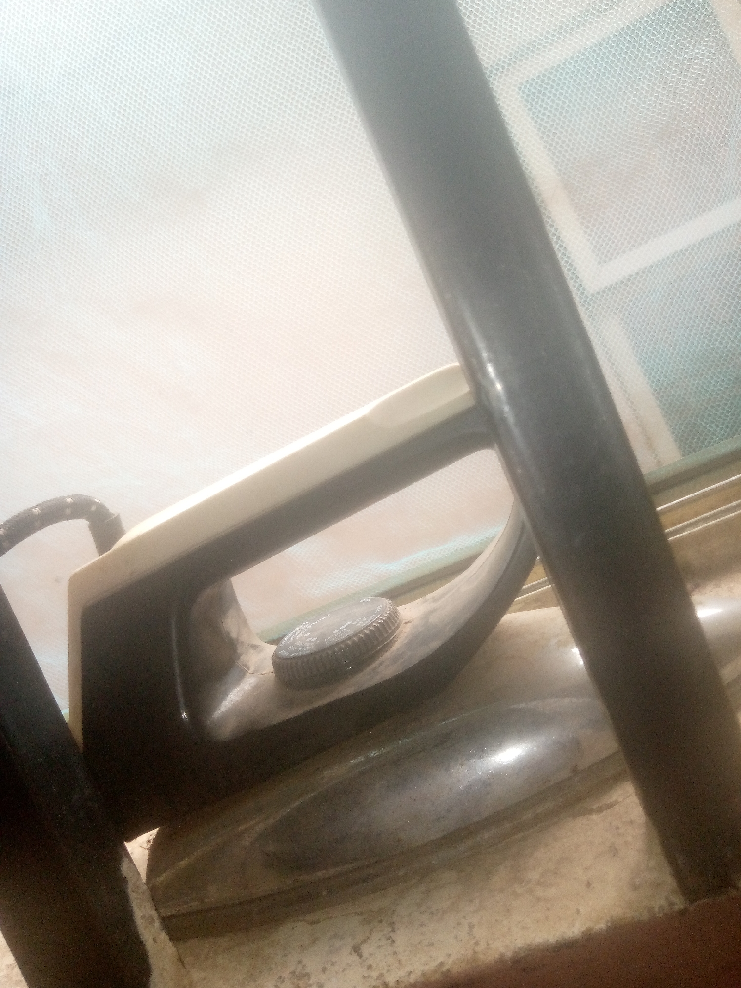 An electric clothes iron rests on a light-colored, textured surface in a setting identified as Unknown, Unknown. The iron features a dark-colored body and a lighter, off-white handle. A circular temperature control dial, with faint markings, is visible on the top of the iron's body. A braided power cord exits the left side of the iron's base. Two dark, cylindrical vertical poles partially frame the scene, one on the left and one on the right, obscuring portions of the iron and the background. The background consists of a finely woven, light-colored mesh material, likely a net or screen, exhibiting a distinct grid pattern. Behind the mesh, there are faint indications of a rectangular structure, possibly a window frame, against a subtly bluish-green backdrop. The lighting appears bright, causing some overexposure in the upper background areas. The overall scene is static, devoid of people or discernible actions.