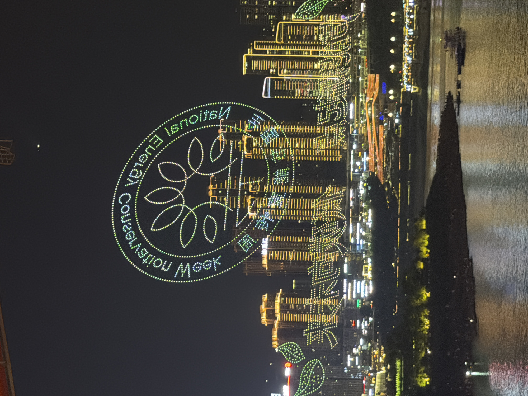 The image captures a vibrant nighttime scene in the Yuzhong District of China, likely a waterfront area illuminated by a stunning display of city lights and festive decorations. The primary focus is a large, circular LED light display that forms a prominent graphic. Within this circle, text is visible, reading "National Energy Conservation Week" in English and possibly Chinese characters. The text is arranged in a curved fashion, following the perimeter of the circle, with some characters appearing in green and others in white.
Surrounding the central light display are numerous tall buildings, their facades covered in intricate patterns of bright yellow and white lights, creating a dazzling urban landscape. These lights suggest a celebration or event, possibly related to the "Energy Conservation Week" advertised. The buildings are reflected in the water, which occupies the right side of the frame, adding to the visual spectacle. The water's surface is textured and reflects the city lights, creating a shimmering effect.
A dark silhouette of a tree or structure juts out from the right side of the frame into the water. In the distance, on the water, a small boat with lights can be seen, suggesting movement and activity even at night. The overall atmosphere is one of festivity and urban grandeur, with the extensive use of lights highlighting the modernity and energy of the city. The time of day is clearly night, with the darkness of the sky contrasting sharply with the brilliance of the illuminations. The weather appears to be clear, allowing for unobstructed views of the light displays.