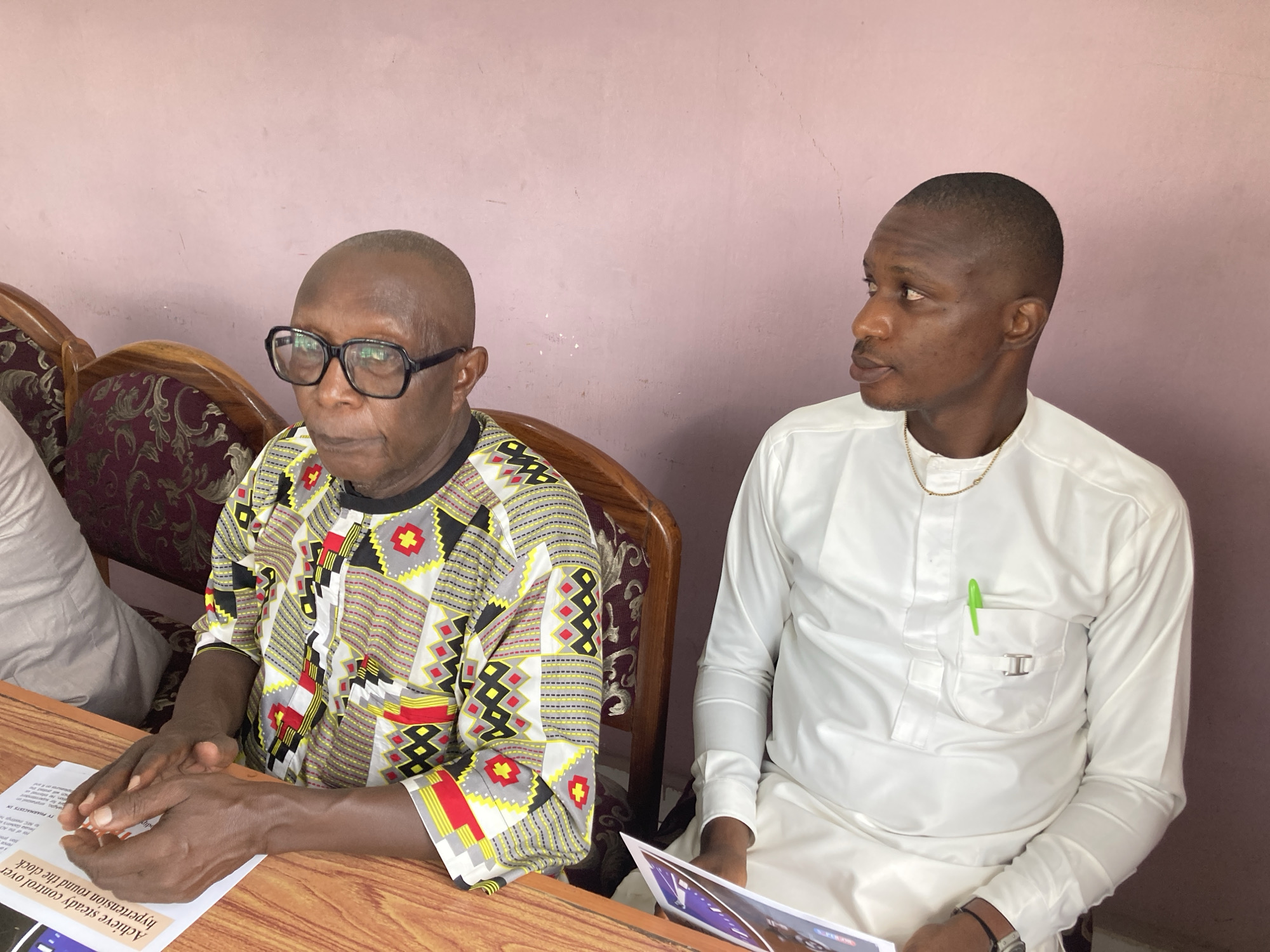 Two men are seated at a wooden table in an indoor setting, likely a meeting or discussion. The man on the left is older, bald, and wears black-framed glasses. He is dressed in a vibrant, multi-colored African print shirt featuring patterns in black, white, red, yellow, and grey. His hands are clasped over a document placed on the table in front of him, and he has a serious or contemplative expression.
To his right, a younger man with a short, neat haircut is wearing a white traditional long-sleeved outfit, possibly an agbada or similar style, adorned with a gold chain around his neck. A bright green pen is clipped into his left breast pocket. He holds a brochure or pamphlet in his hands and is looking intently towards the older man, suggesting he is listening or engaged in a conversation. Part of a third person in a light-colored top is partially visible on the far left.
The background features a plain light pink wall. The men are seated in wooden chairs with ornate, carved backs and dark maroon patterned upholstery. On the document in front of the older man, visible text reads: "Achieve steady control over hypertension round the clock" and "Pharmagists," indicating a health-related topic. The overall atmosphere appears formal and focused.
