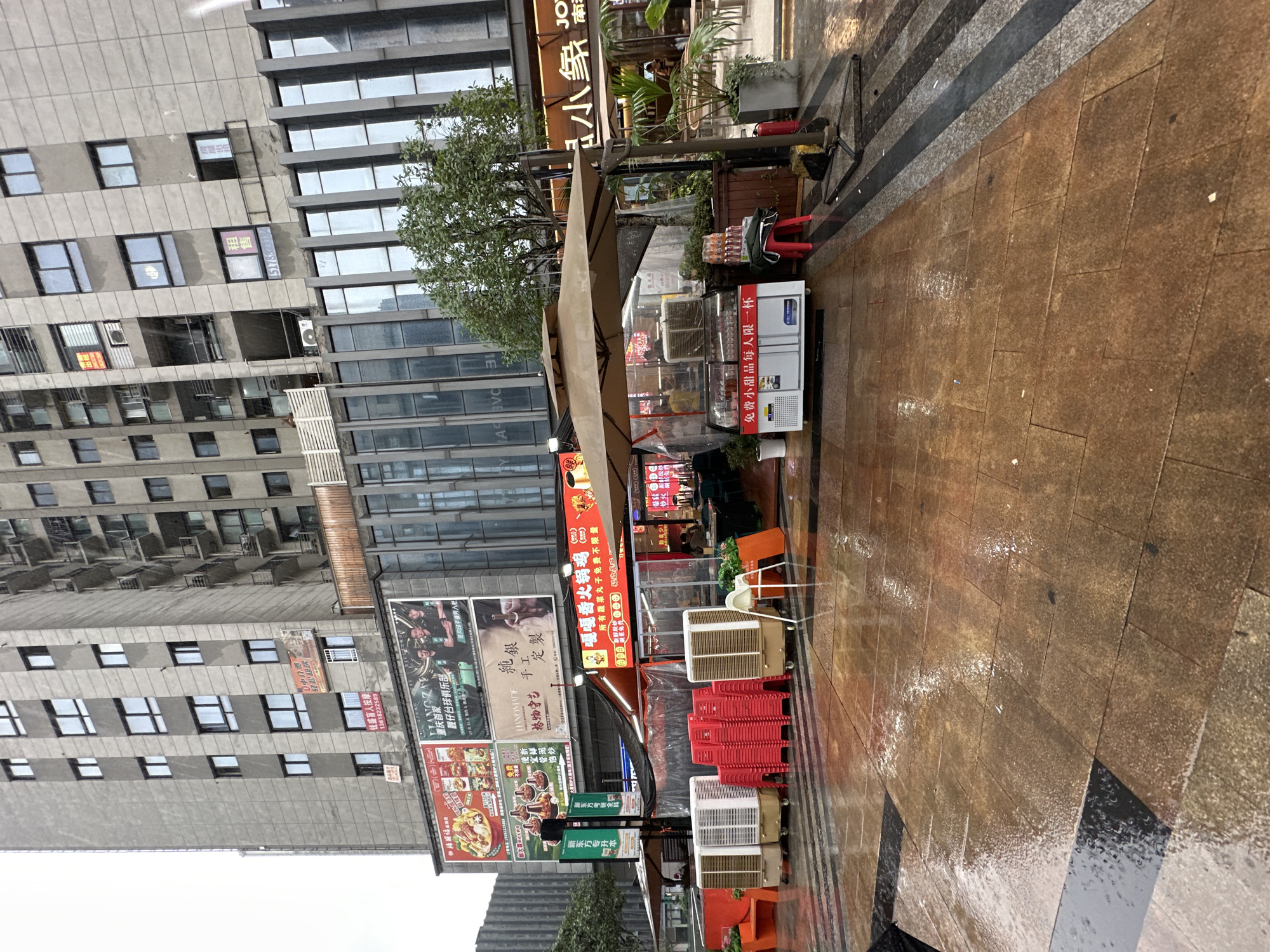 This detailed description pertains to an outdoor commercial scene in Chongqing High-tech Zone, China, following a period of rain, evidenced by the wet pavement. The image is dominated by a tall, multi-story, grey concrete building that appears to be residential or mixed-use. It features numerous rectangular windows, many of which are dark, though some show interior lights or reflections. The right side of the building reveals several balconies, some with visible items. Below the residential section, a commercial facade extends, characterized by large glass panels with dark vertical frames. On the commercial facade, several large billboards are prominently displayed. The leftmost billboard advertises various food dishes, including hot pot, with the text "福临佳肴" (Fulin Jiayao - auspicious dishes/delicacies) and a phone number (138162232588). The central billboard features three men and the text "LIANGZAI 重庆仔俱乐部" (Liangzai Chongqing Boy Club). The rightmost billboard advertises "HANDMADE 纯银手工制" (Handmade Pure Silver Handmade). Smaller red signs are affixed to the grey building walls; one higher up states "0中介费 轻松找好房" (No intermediary fee, easily find a good house), and another lower down reads "租售 13618252589" (For Rent/Sale, with a phone number). Another "租售" (For Rent/Sale) sign with a partial phone number (5178xxxx) is visible on the right side of the building. At street level, the scene transitions to an active commercial area. A prominent red awning shelters an outdoor dining establishment identified by the text "嘿喝番茄火锅" (Hey He Tomato Hot Pot). A sign below the hot pot name states "所有蔬菜丸子免费不限量" (All vegetables and meatballs are free and unlimited) and "欢迎品尝" (Welcome to taste). Inside this semi-enclosed dining area, tables and chairs are visible, with a few patrons seated. Two large, beige outdoor umbrellas provide additional cover. Along the edge of the paved area, several large, portable evaporative coolers (three beige, one white) are lined up, and another smaller one is within the dining area. A substantial stack of red plastic chairs is positioned between the coolers. An orange planter or bench is visible to the left. A white refrigerated display unit on wheels, containing drinks and small desserts, stands near the right umbrella. A sign on this unit reads "免费小甜品每人一杯" (Free small dessert, one cup per person). A red plastic stool with a stack of bottled/canned drinks rests next to the cooler. A red fire extinguisher is located at the base of one umbrella stand. To the far right, another restaurant, partially visible, has a sign
