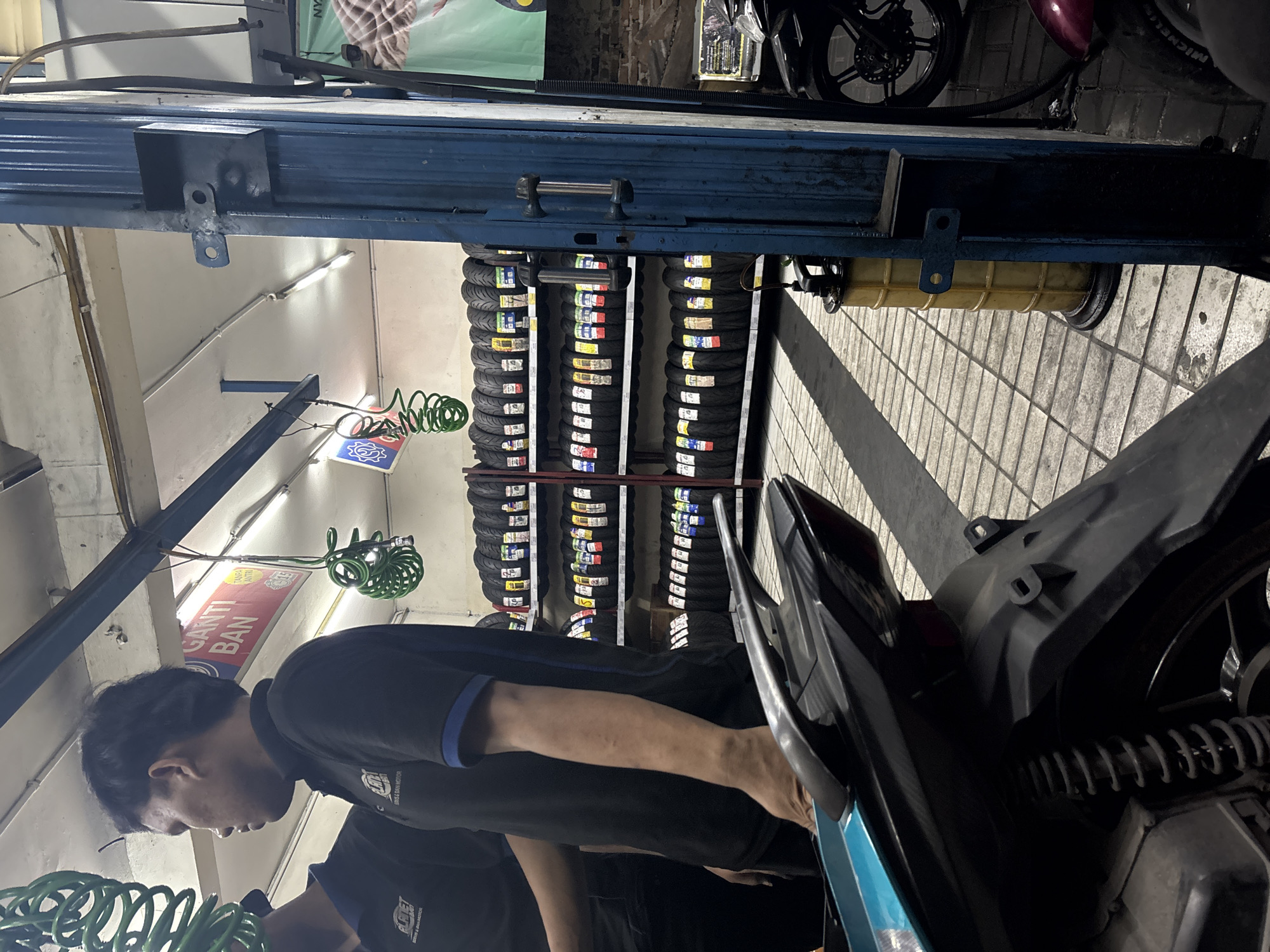 The image depicts the interior of a tire shop located in North Jakarta, Indonesia. In the foreground, the rear of a teal and black motorcycle is visible. To the left, two men in black uniforms stand. One man, in the mid-ground, faces left in profile and appears to be interacting with the other man, whose upper body is visible. Behind the men, shelves are stacked with numerous motorcycle tires. The shop is well-lit with fluorescent ceiling lights. Various tools and equipment, including a green coiled hose and a yellow oil collection drum, are present. A blue metal door frame with handles is on the right side of the image, and part of a black motorcycle is visible to the far right. The floor is tiled with a pattern of grey and white squares. A red banner with white text reads "GANTI BAN" which translates to "CHANGE TIRE".
