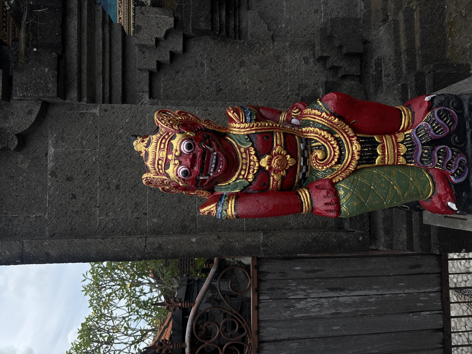 The image displays a brightly colored, carved statue positioned horizontally in the frame. The central subject is a traditional guardian figure, predominantly red, with intricate gold, green, and blue detailing. The figure has a fierce facial expression, characterized by wide, staring white eyes, prominent teeth, and a decorative golden crown or headdress. Its body is adorned with swirling gold patterns, and it wears a green lower garment, with large red hands.
Behind the statue, dark gray, textured stone architecture forms the background, featuring carved blocks and what appear to be steps or decorative patterns. A substantial horizontal stone beam or archway runs across the top of the frame. In the bottom left corner, green foliage from a tree is visible against a light sky, indicating daytime. The bottom right shows a dark wooden plank structure, possibly a gate or wall, alongside a section of a rusty wrought-iron railing and a light-colored pebbled ground. No specific activity is depicted, as the scene is static, showcasing the cultural artifact within its architectural setting. No text is visible in the image. The statue's style is characteristic of Balinese guardian figures found in temples or traditional entrances, consistent with the North Kuta, Indonesia context.