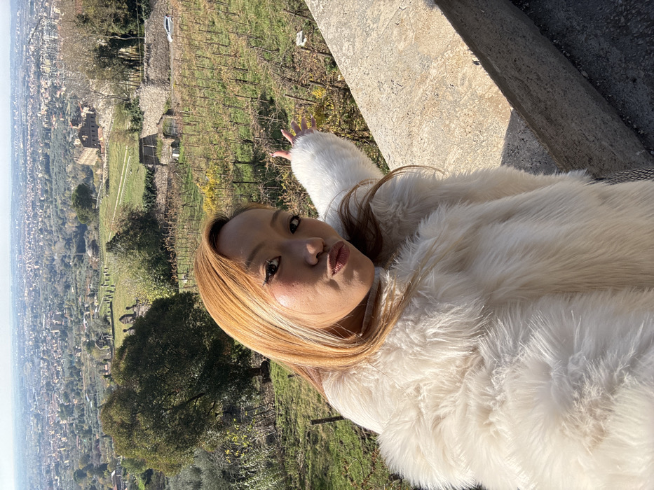 The image displays a panoramic landscape captured from an elevated position, with a female individual in the foreground. The woman, of apparent East Asian ethnicity, is positioned slightly left of the frame's center, wearing a cream-colored faux fur coat and exhibiting light brown hair with blonde highlights. Her face is directed towards the camera with puckered lips and visible eye makeup. Her right arm is raised with an open hand, pointing towards the background scenery. She stands beside a light-colored, textured concrete or stone railing, which diagonally traverses the lower right quadrant of the image.
The background reveals a sunlit valley under a clear blue sky, characteristic of Tivoli, Italy. Immediately beyond the railing and sloping downwards is a vineyard, featuring rows of grapevines with minimal foliage, suggesting a late autumn or winter season. Scattered green trees are interspersed throughout the landscape. In the mid-ground, extensive ancient Roman ruins, consistent with Hadrian's Villa (Villa Adriana), are clearly visible, presenting large brick structures and segmented walls. A white vehicle is present on a road winding through this area. The far background encompasses a sprawling urban expanse with numerous buildings, indicative of the city of Tivoli and its surrounding regions, culminating in distant hills or mountains on the horizon. Bright sunlight illuminates the scene, casting distinct shadows.