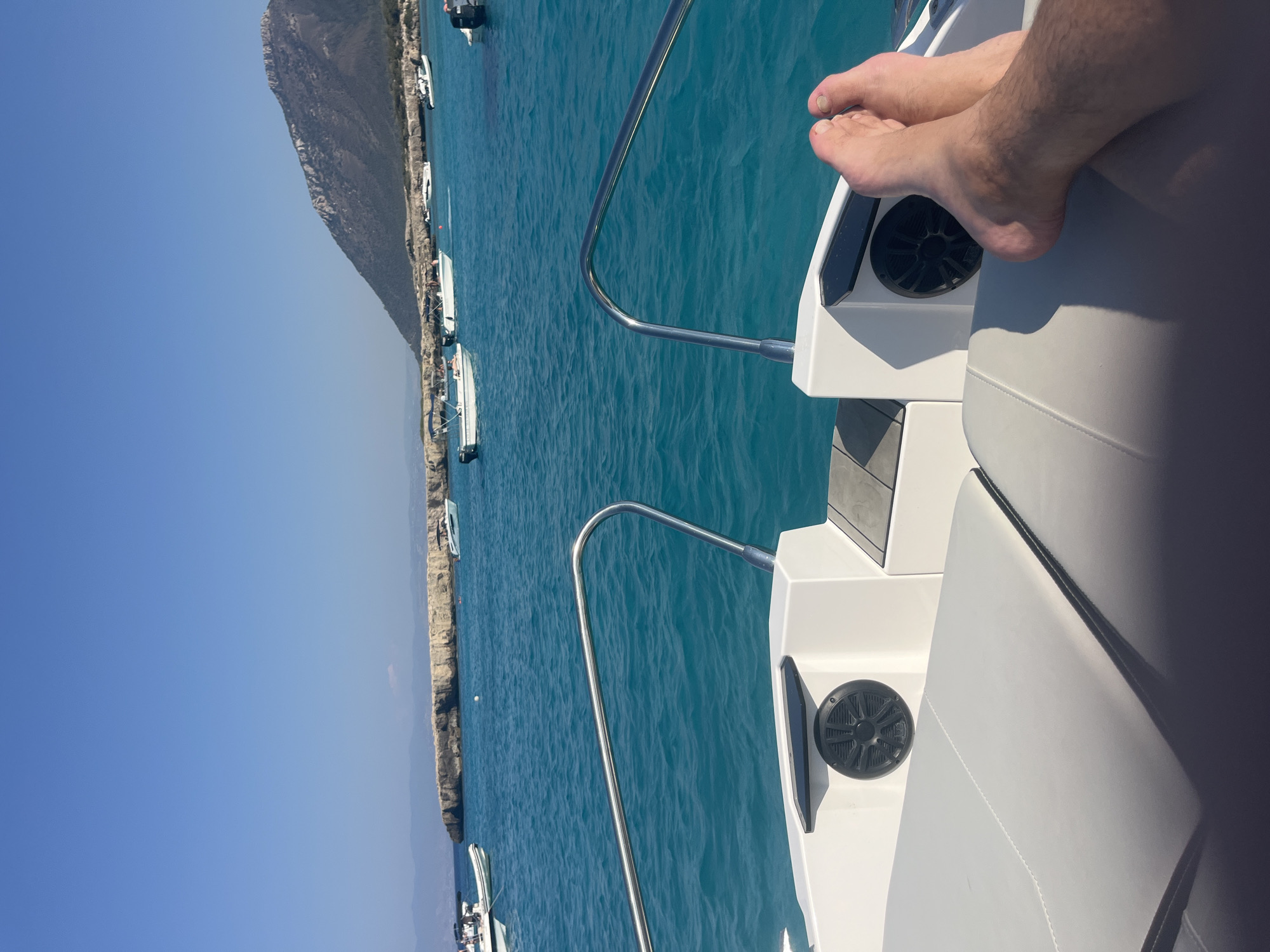 The image captures a point-of-view shot from a boat on a clear, sunny day. The foreground shows the deck of the boat with grey cushioned seating and a person's bare feet resting on it. A chrome railing is visible to the left and right. In the mid-ground, the turquoise waters of the sea are calm with several other boats scattered across the surface. In the background, a rocky coastline is visible with a large, mountainous terrain rising behind it. The sky is a vibrant, cloudless blue. This scene is consistent with locations around Neo Chorio, Cyprus, known for its beautiful coastline and clear waters which attract boating activities.