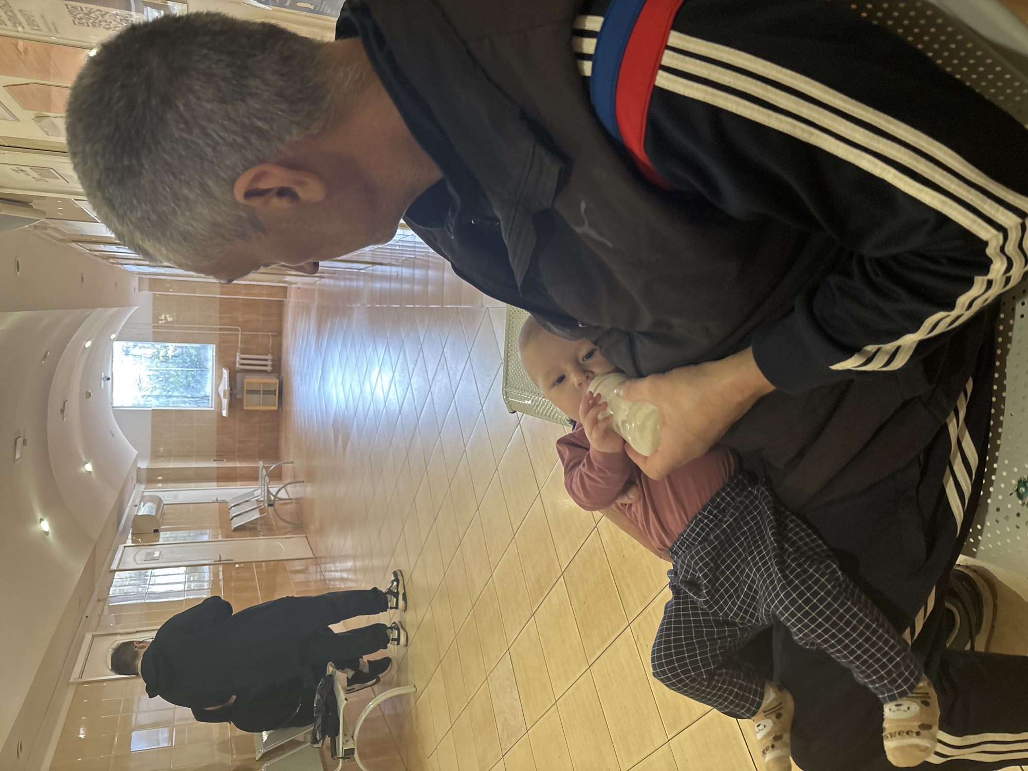In what appears to be a hospital or clinic waiting area, an older man with short gray hair is seated and holding a baby who is drinking from a bottle. The man is wearing a black jacket with white stripes on the sleeve and a blue and red stripe across the back. The baby is wearing a pink shirt and blue checkered pants. In the background, there are rows of chairs and a person walking away. The location is likely in Sochi, Russia, based on the potential reference of the blue, red, and white colors on the jacket matching the colors of the Russian flag.