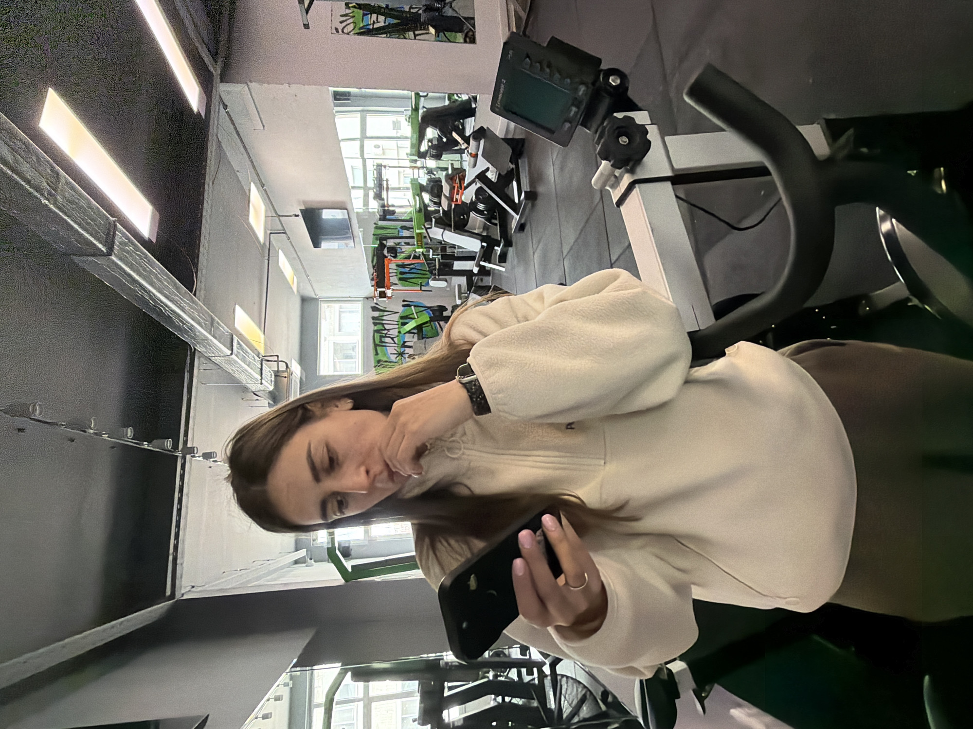 A woman stands in a gym holding a black smartphone. She is wearing a cream-colored fleece pullover, and a ring on her finger and a smartwatch on her wrist are visible. The gym has various exercise machines in the background, including rowing machines and other equipment. The environment appears to be a modern gym, possibly in Sofia, Bulgaria.