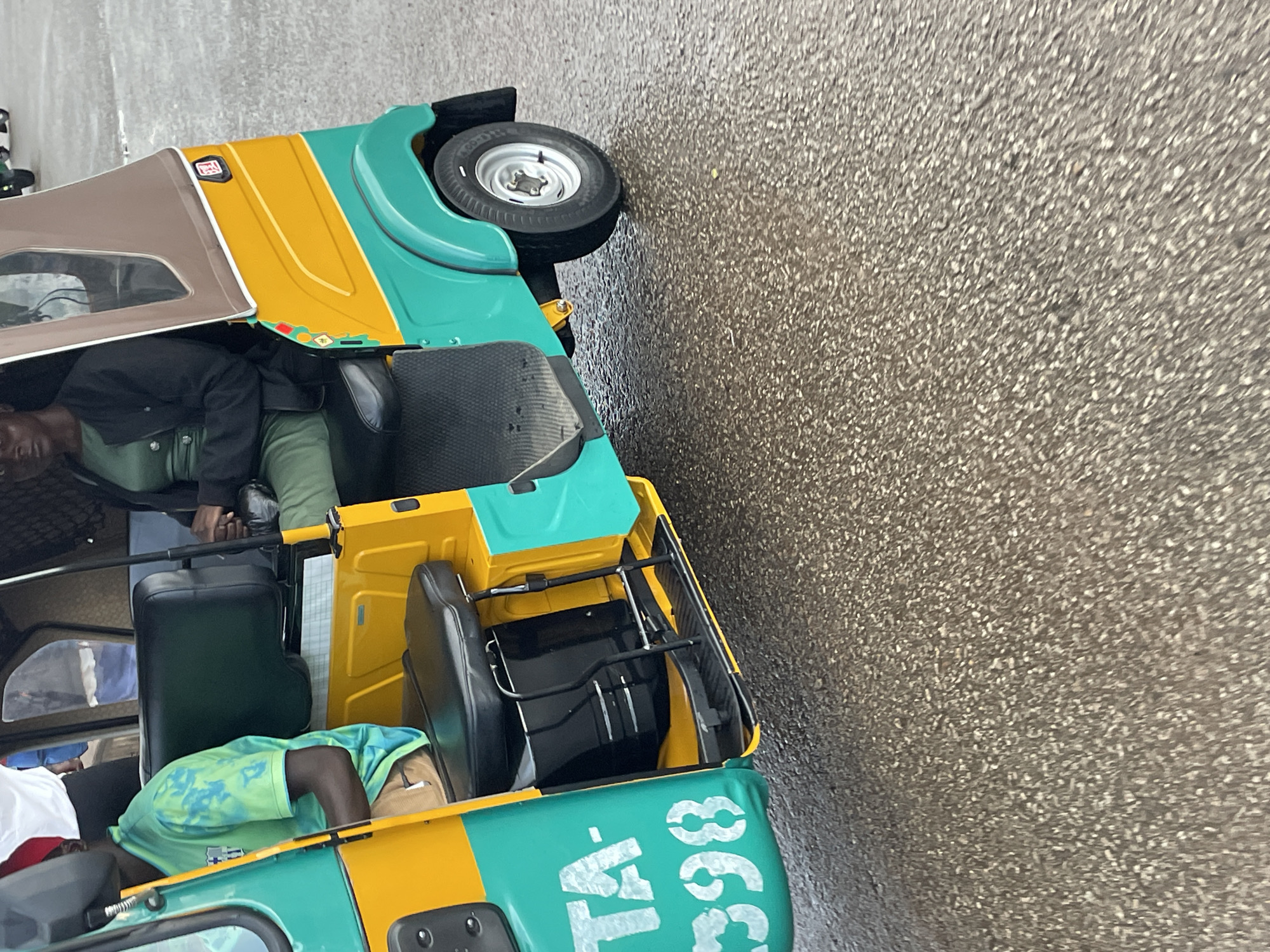 An outdoor, eye-level, medium shot captures a three-wheeled auto-rickshaw in Barakin Lerawi, Nigeria. The auto-rickshaw is painted in vibrant yellow and teal, with a brown canopy. A Black male, wearing a green tie-dye shirt and tan shorts, sits in the front seat, likely as the driver. Beside him, a Black female, dressed in a green dress and black jacket, is seated in the passenger area. The vehicle appears to be stationary on a wet, asphalt surface, suggesting recent rainfall. White lettering on the side of the auto-rickshaw reads "TA-398". The surrounding environment is not clearly visible, but the pavement texture indicates it is an urban area.