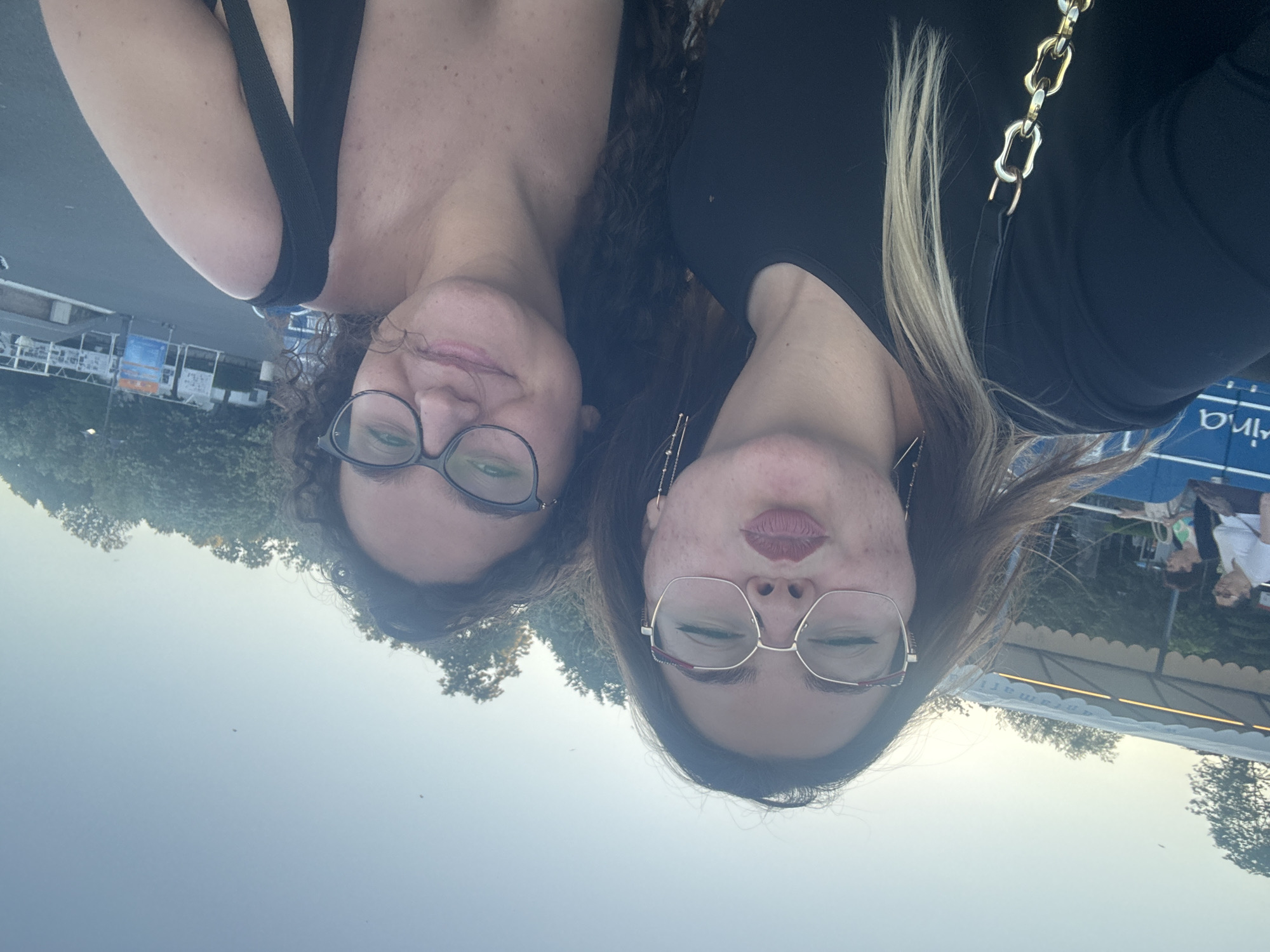 Two young women are captured in a selfie outdoors with a clear sky overhead. The woman on the left, wearing glasses and a black top, puckers her lips. Her hair is dark with lighter streaks. She has a gold chain bag strap visible. The woman on the right, also wearing glasses, smiles at the camera. Her dark, curly hair frames her face. In the background, there are trees and structures suggesting an outdoor venue, possibly a waterfront promenade or park. Faint blue and white signage is visible in the middle distance. The lighting indicates it is likely late afternoon or early evening. Based on the signage with the word "Marina" visible on the blue structure behind the woman on the left, this scene is likely in the vicinity of the Marina in Burgas, Bulgaria, a popular coastal city known for its seaside attractions.
