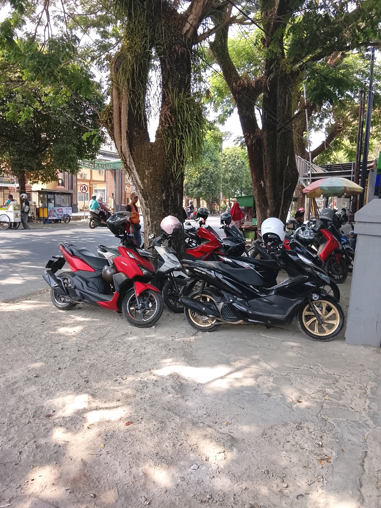 The image depicts a street scene in Slawi, Indonesia, specifically in front of a building that appears to be a school, indicated by the sign "NEGERI 2 TEGAL" (State Junior High School 2 Tegal). Several motorbikes are parked along the side of the road. A red and black motorbike is in the foreground on the left, followed by a black motorbike with gold rims, and further back, other motorbikes are visible. A person wearing a white helmet is seated on one of the motorbikes. In the background, a food cart is present, and a few people are walking or riding motorbikes. The environment features large trees providing shade, suggesting a sunny day. The ground is a mixture of asphalt and dirt. A muted colorful umbrella is visible on the right side of the image, possibly associated with a vendor.