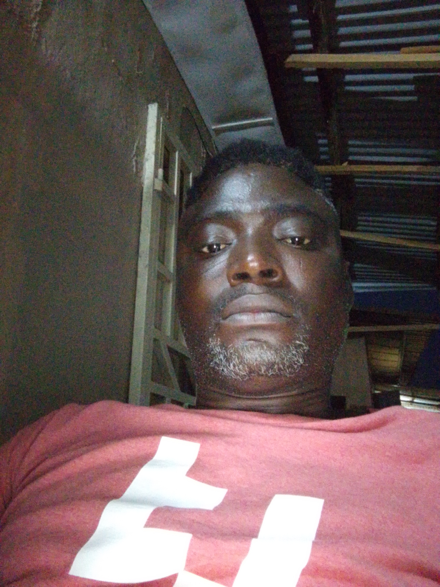 In Sabon Tasha, Nigeria, a man is captured in a close-up, selfie-style shot, appearing to be indoors. He is wearing a reddish-pink t-shirt with a large white graphic design across the chest. The man has dark skin, short, dark hair, and a light-colored beard and stubble. His eyes are open, and he is looking directly at the camera. Behind him, the environment is dimly lit and appears somewhat unkempt. To the left, a dark, textured wall is visible, with a section of a light-colored, possibly metal, frame structure against it. To the right and in the background, a corrugated metal roof is supported by wooden beams, suggesting a rustic or unfinished interior. The lighting suggests the image was taken with a flash.