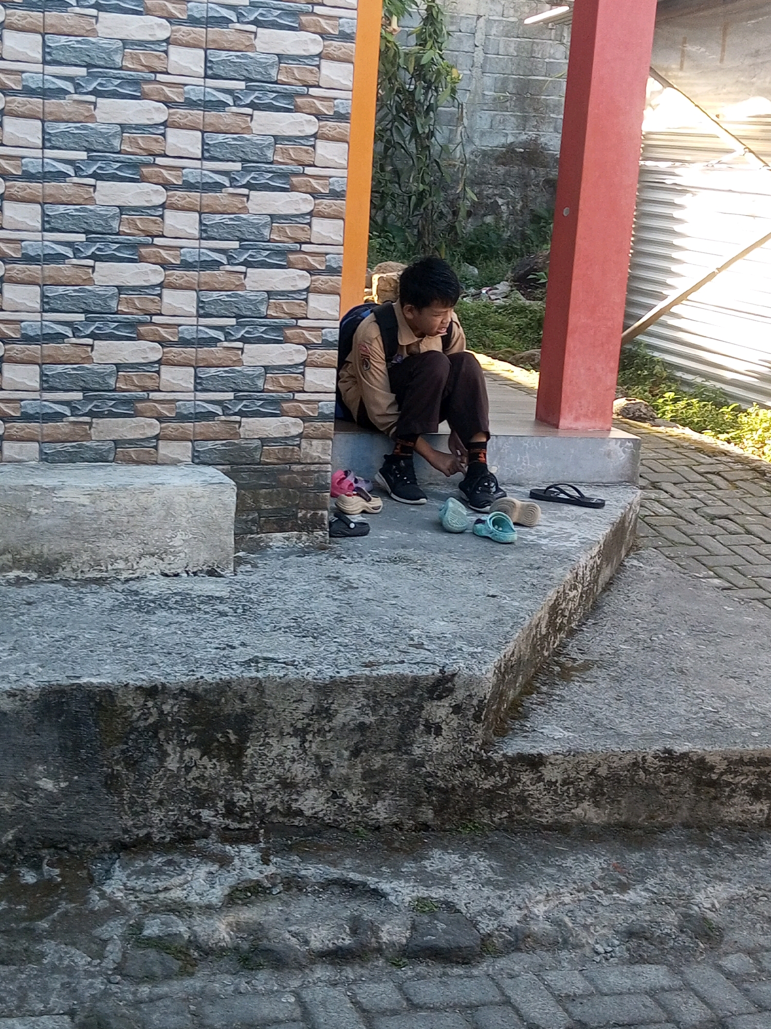 A young boy wearing a school uniform sits on concrete steps outside a building with a tiled facade. He is hunched over, looking down and resting his chin on his hand. Several pairs of footwear are scattered around him on the steps and ground, including sandals and closed-toe shoes. In the background, there is a red pillar, a corrugated metal structure, and overgrown vegetation. The scene appears to be in a public outdoor space, possibly near a school in Andongsili, Indonesia, given the context.