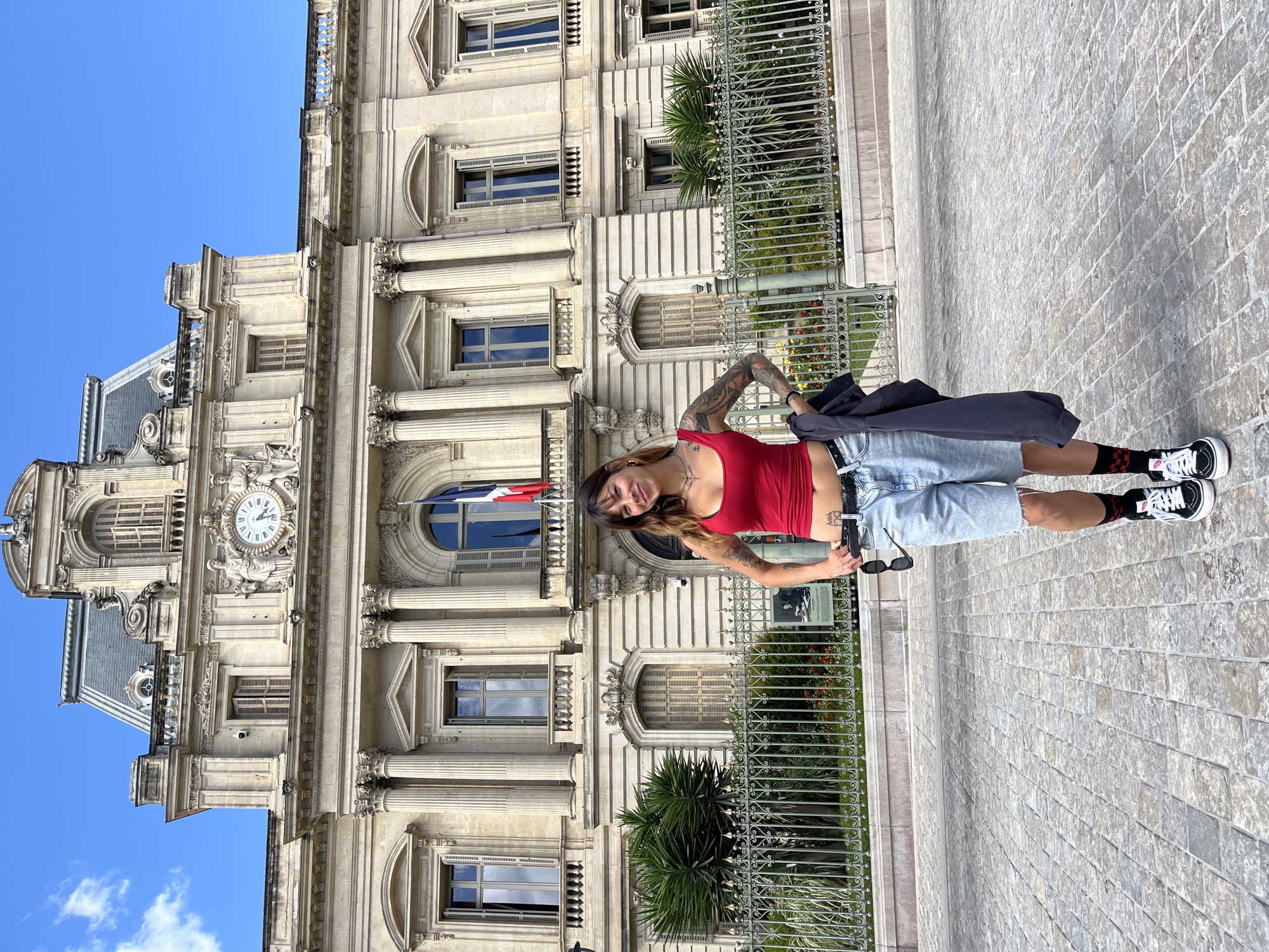 A slightly rotated, full shot shows a young woman standing to the right of a tan, ornate building with columns in Montpellier, France, set against a blue sky.
The woman faces the camera, her head tilted to her left. She has dark hair and fair skin with tattoos visible on her arms. She wears a red tank top, light blue denim shorts, black and red socks, and black and white sneakers. She is holding a pair of sunglasses and a dark jacket.
The building behind her is highly detailed with classical architectural elements, including columns, arched windows, and ornate carvings. A clock is visible on the building's facade. A French flag is flying from the building. A decorative fence separates the sidewalk from the building. The ground is paved with grey stone bricks.