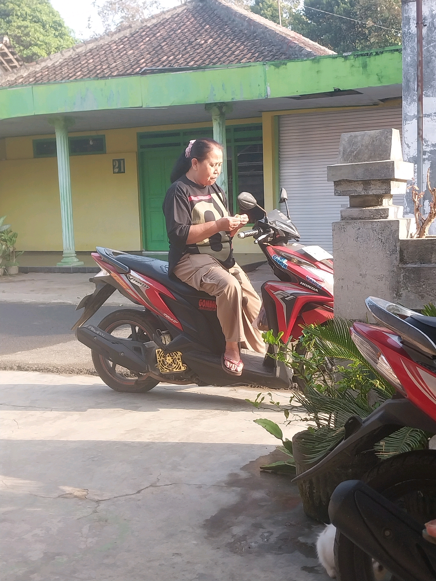 In Nglegok, Indonesia, a woman sits on a red and black scooter in front of a yellow building with a green trim. She is wearing a black t-shirt and tan pants, and her hair is pulled back with a pink scrunchie. The woman is looking down at her hands, which are holding a small object. The building appears to be a house or shop, with a roll-up door and a number "21" visible on the wall. There are trees and other vegetation around the building, and the sky is visible in the background. Another scooter, partially visible, is parked to the right of the woman's scooter. The scene is well-lit by sunlight.