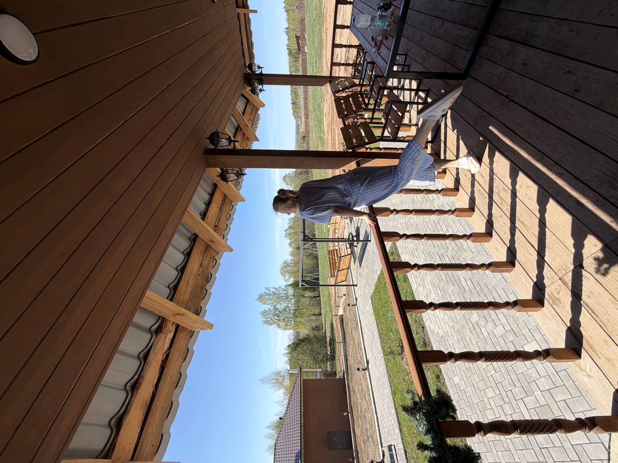 A woman wearing a blue and white striped dress and white sneakers stands on a wooden porch. She is grasping a wooden handrail of stairs. The scene is outside a wooden building, likely a house or cabin. The sky is blue, and trees and green fields are visible in the background. A table and chairs are also visible on the porch. The ground is covered in gray paving stones. The location is Kamennoye, Russia.