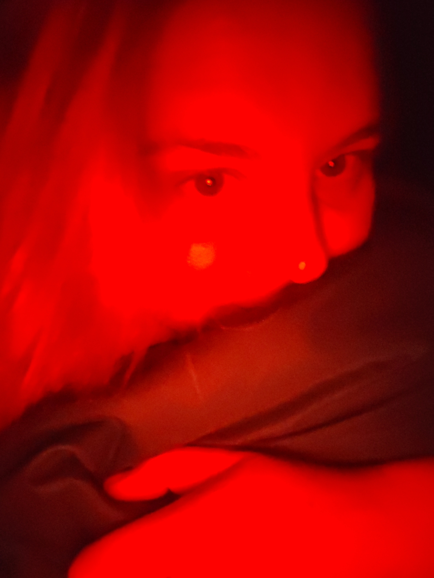 A person's face is illuminated with red light. The individual has fair skin, dark eyes, and hair that is blurred from the red light. The person appears to be partially obscured by or holding a dark-colored fabric or garment close to their face. No information suggests any connection to Izhevsk, Russia.