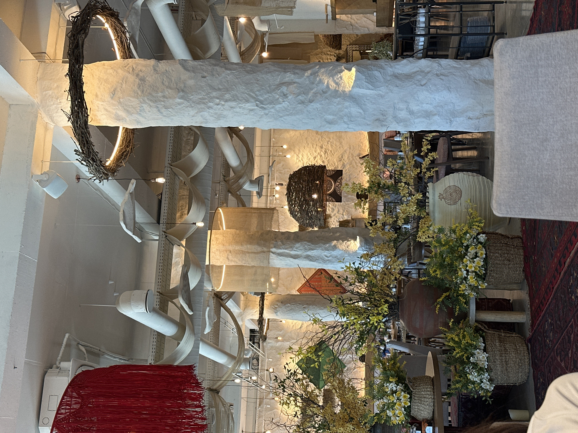 This image depicts a warmly lit interior space, likely a restaurant, cafe, or shop, characterized by an eclectic and rustic design aesthetic. The photo is oriented vertically, with the scene appearing on its side.
The prominent feature is a long, textured, off-white beam or architectural element running horizontally across the middle of the frame (when viewed upright). Above this beam, the ceiling is adorned with white pipes, flowing fabric elements resembling scrolls or ribbons, and numerous exposed bulb light fixtures. A striking circular light fixture made of natural branches, incorporating a warm LED light strip, hangs prominently. Further to the left, a vibrant red fringed textile or artwork adds a bold splash of color.
Below the central beam, the area appears to be a seating or dining space with tables and chairs partially visible in the background. Abundant decorative plants with yellow and white flowers are arranged in large terracotta-colored pots and woven baskets, contributing to a natural, earthy feel. One pot features a distinct pomegranate-like design. A dark red patterned rug with geometric motifs covers a portion of the floor on the right. Another chandelier, crafted from dark branches, hangs deeper in the room. The walls are textured, possibly stucco or rough plaster, complementing the rustic theme.
No specific human activity is visible, but the arrangement suggests a setting for dining or social gathering. The lighting is predominantly warm and ambient, enhancing the inviting atmosphere. The design emphasizes natural materials, varied textures, and unique lighting fixtures, creating an organic and artisanal ambiance. No discernible text is visible in the image. The scene is likely illuminated by artificial light, suggesting evening or an interior space.