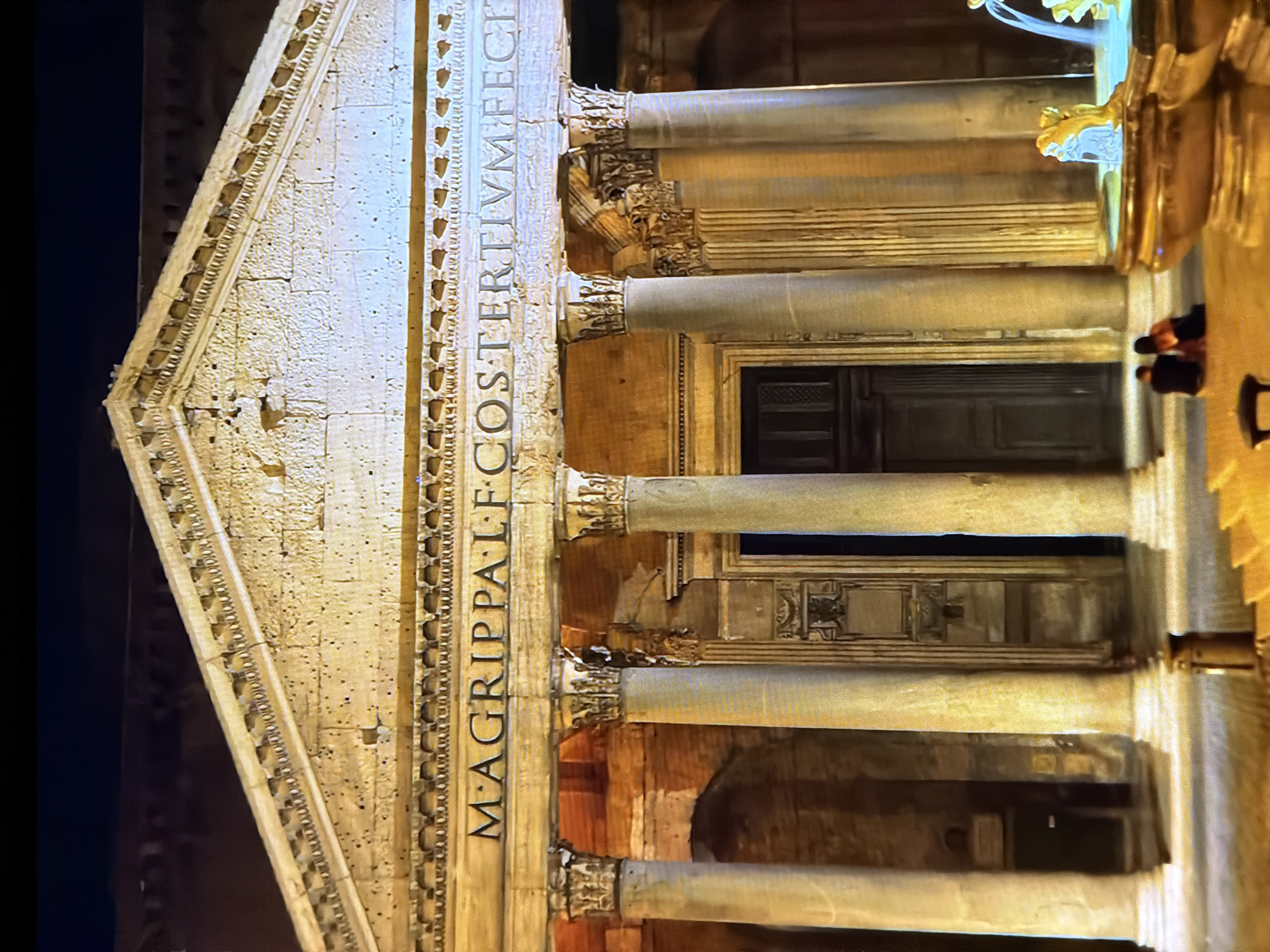 The image shows the exterior of a large, ancient building with classical architecture at night. The building features a pediment with decorative carvings and an inscription that reads "M. AGRIPPA L. F. COSTERVIM FECIT". It is supported by massive columns. In front of the building, there are steps leading up to the entrance. To the right, a fountain with sculptures is visible. Two people are seated on the steps, facing away from the camera, appearing to be a couple. The lighting suggests it is evening or nighttime, with artificial lights illuminating the facade. The city of Chirchik, Uzbekistan is not depicted in this image.