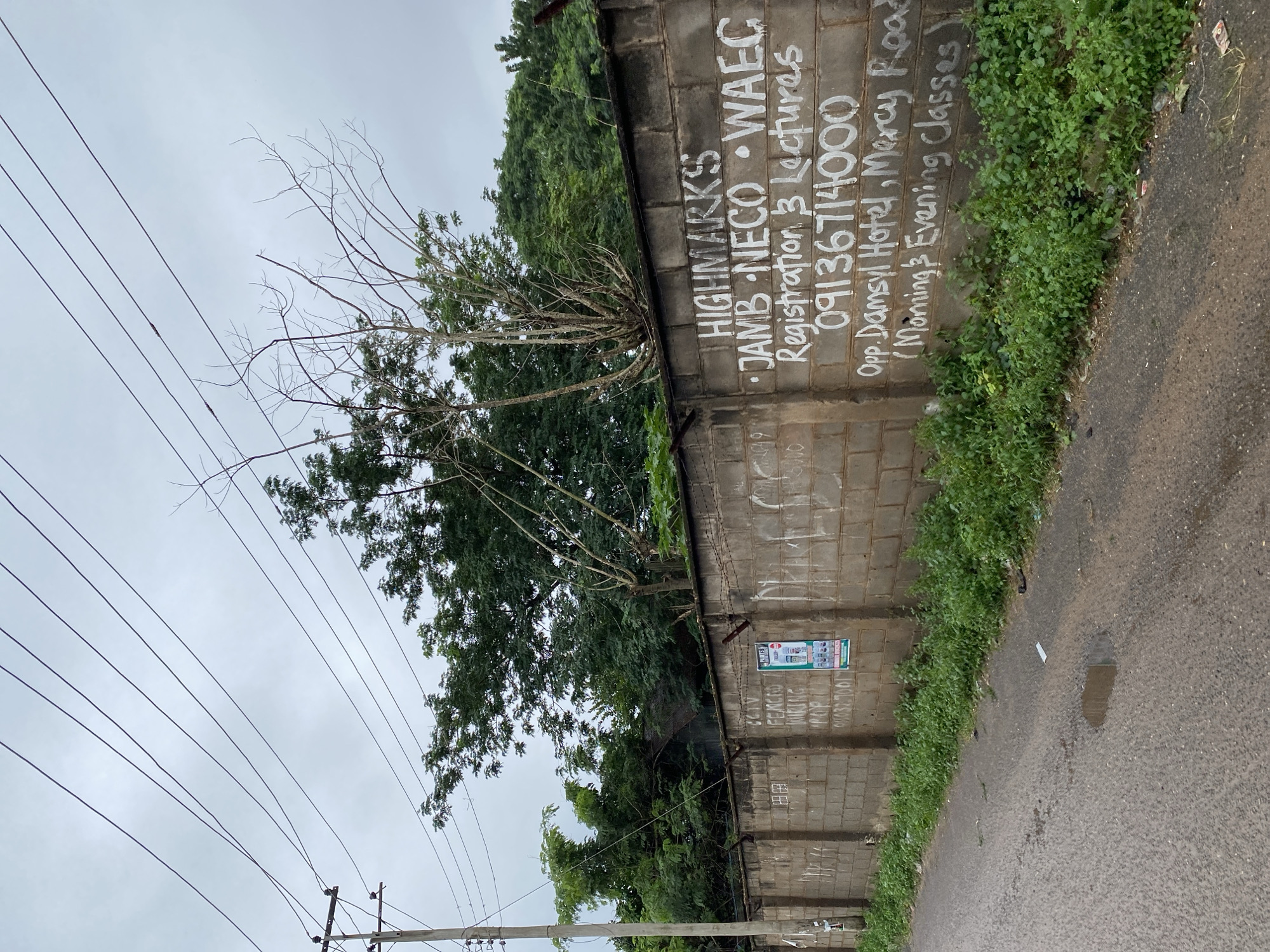 The image depicts a concrete block wall in Abeokuta, Nigeria, with various writings and a poster. The wall appears to be a boundary for a property and is topped with barbed wire. On the wall, in white spray paint, are the words "HIGHMARKS JAMB NECO WAEC Registration & Lectures 09136714000 Opp. Damsyl Hotel, Mercy Road (Morning & Evening Classes)". A poster is also affixed to the wall, featuring images of paint cans. To the left of this writing, some less legible text is present, including what appears to be "CCTV FENCES WUKING POR". The wall is adjacent to a road with some patches of wetness and vegetation growing along its base. In the background, trees and a utility pole with overhead wires are visible against a cloudy sky.