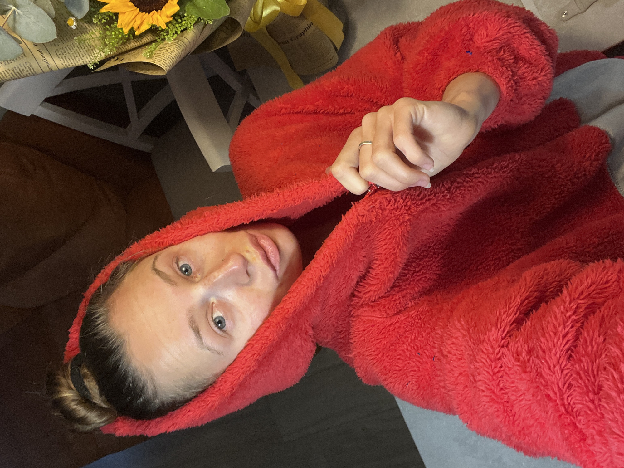 This image depicts a female subject in a close-up, selfie-style indoor photograph taken in Bečići, Montenegro. The subject, an adult female, has light skin with visible freckles, blue eyes, and her hair is pulled back into a bun secured with a black elastic. She has a small stud piercing on the right side of her nose and two small studs near the corner of her left eye. She is wearing a vibrant red, fuzzy, hooded jacket with a shaggy texture, and a light grey garment is visible underneath. Her right hand, wearing a plain silver-colored ring on the ring finger, is holding the zipper pull of her red jacket. She is looking directly towards the camera with a neutral expression. In the background, to the subject's right, a bouquet of flowers is present, featuring a prominent sunflower, smaller yellow flowers, and green foliage, wrapped in newspaper-like paper with a yellow ribbon. Partially visible behind the subject is dark orange or brown upholstered furniture on the left and a white chair with a slatted backrest on the right. A light grey surface, possibly a tabletop or counter, is visible in the lower right foreground. The scene is illuminated by artificial indoor lighting.