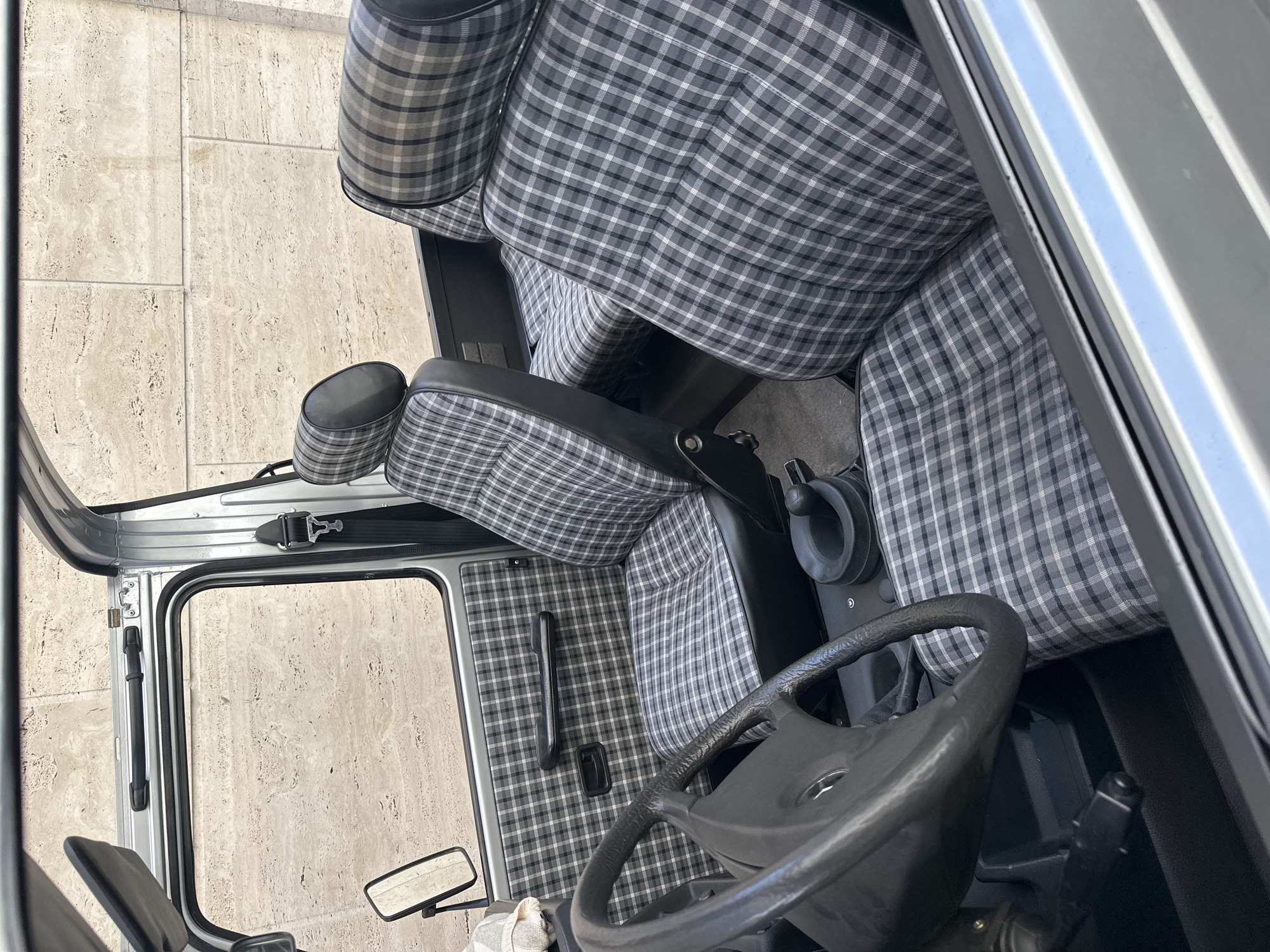 The image shows the interior of a vehicle, with a focus on the driver's seat, steering wheel, and door. The upholstery of the seats and door panels features a gray and white plaid pattern. The steering wheel is black and has a Mercedes-Benz logo in the center. The door is open, revealing the exterior of the vehicle. The image is rotated 90 degrees clockwise. The exact location in Milan, Italy is not specified or implied by the image.