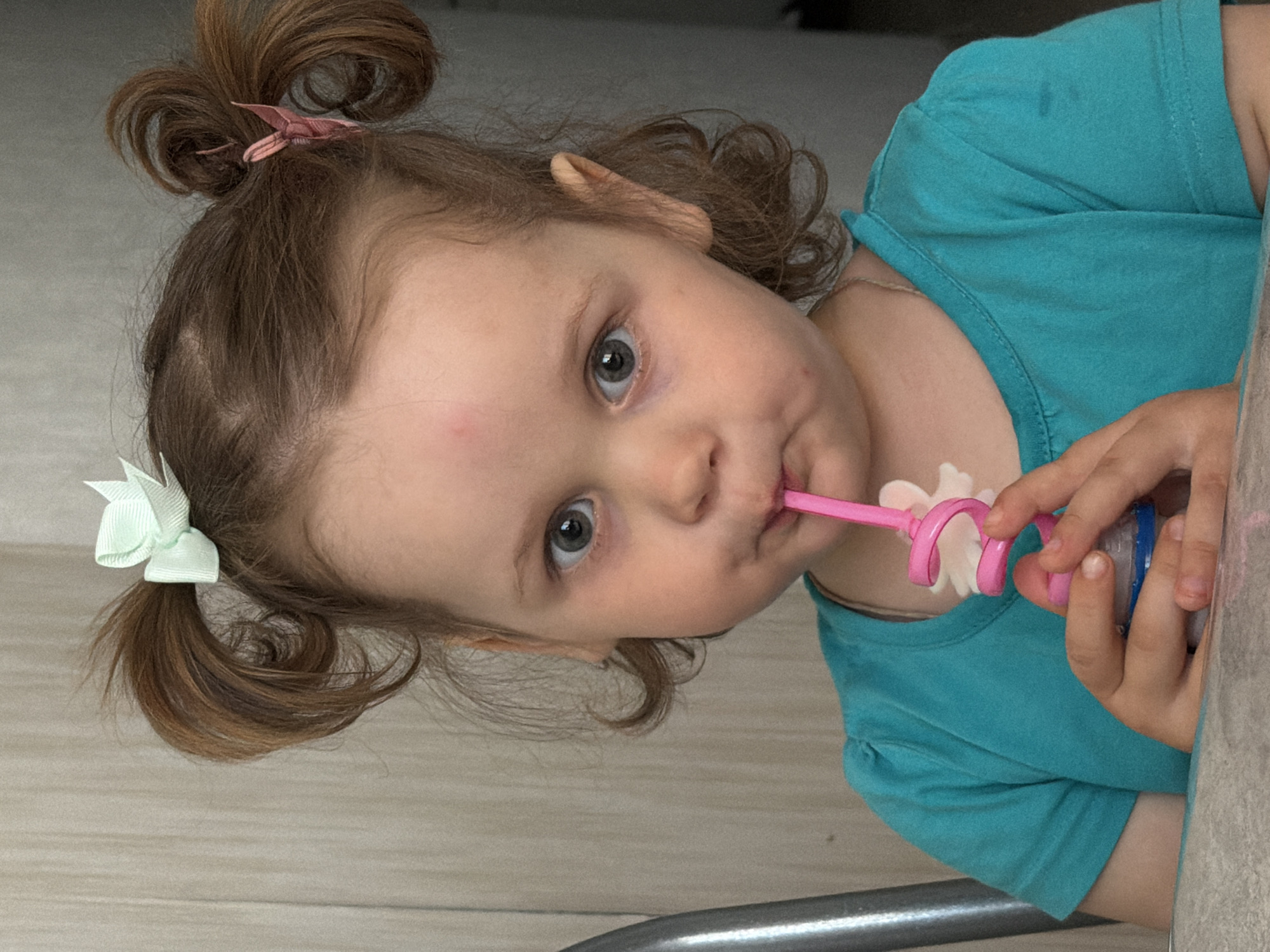 A young child, likely a toddler, is captured in a close-up, tilted portrait indoors. The child looks directly at the camera with attentive, wide blue-grey eyes and a focused expression. They have light brown hair styled in two pigtail buns; one is secured with a small pink clip, and the other with a light green bow. The child's fair skin shows a few faint red marks on the forehead and cheek.
The child is wearing a short-sleeved turquoise t-shirt, which has minor smudges on the upper left shoulder. They are actively using a feeding device, holding a blue and clear plastic container with both hands. A flexible pink, coiled straw-like component, featuring a white flower-shaped accessory, extends from the container into the child's mouth, indicating they are consuming something.
The background is simple, consisting of light-toned surfaces, possibly wood paneling or light-colored walls, consistent with an indoor domestic environment. A metallic rail or bar is visible in the lower part of the frame, suggesting the child might be in a high chair or seated near a piece of furniture. The overall impression is a candid moment of the child during a meal or snack time in a home setting in Chaikovsky, Russia. No visible text is present in the image.