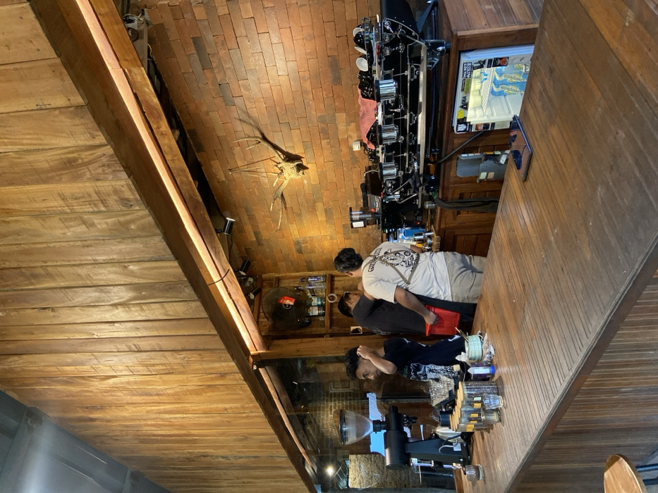 The image depicts the interior of a cafe or coffee shop located in Cembor, Indonesia. The scene is dominated by a rustic aesthetic, featuring a long, dark wooden counter in the foreground with horizontal grooves on its top surface. On the counter, various small objects are arranged, including several glass jars with lids, glasses, and a blue container. A smartphone rests flat on the counter toward the right.

Behind the counter, a service area is visible with a brick wall backdrop. The upper wall sections are clad in vertical wooden planks. A prominent wooden beam with integrated strip lighting spans horizontally, separating the wooden and brick wall sections. Mounted on the brick wall are a large pair of deer antlers. To the left, a metal fan is affixed above shelves stocked with bottles.

Three adult males are positioned behind the counter. The leftmost male, with dark curly hair, faces right while looking forward, wearing a dark t-shirt. The central male is partially obscured, facing right, and holds a red plastic basket. The rightmost male has his back to the camera, wearing a light-colored t-shirt with a graphic print and light-colored pants. He is standing near a large, professional black espresso machine with multiple group heads, on top of which several cups are inverted. To the left of the espresso machine, a black commercial coffee grinder with a transparent bean hopper is visible. Below the espresso machine, a small white refrigerator containing bottled beverages is integrated into the counter structure. The individuals appear to be staff members engaged in service activities.