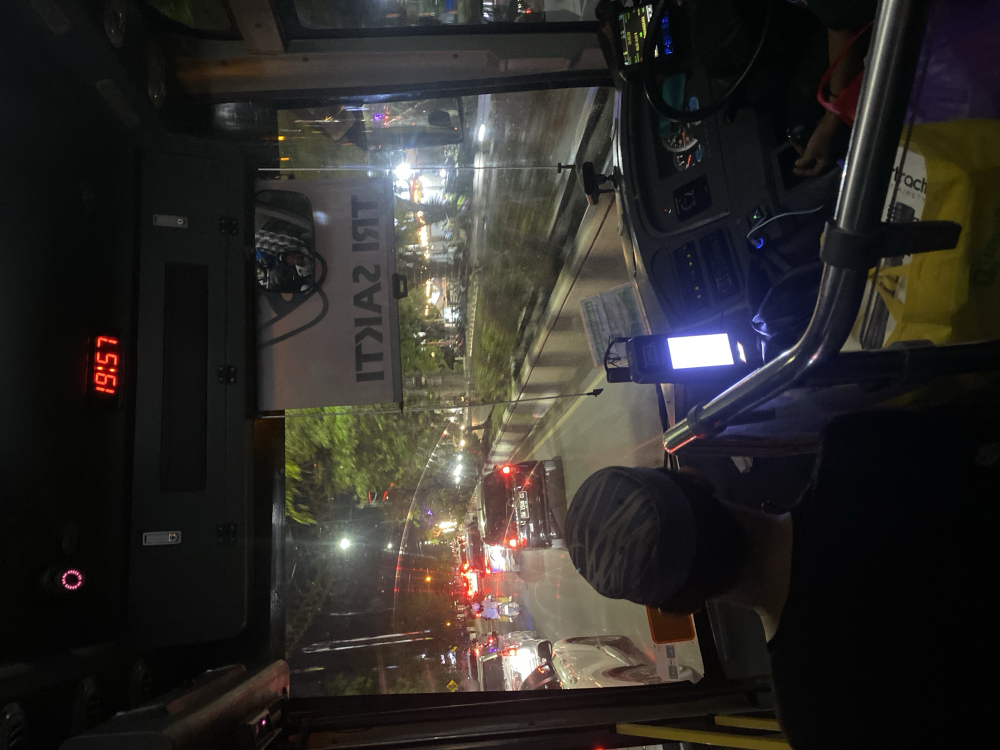 The image captures the interior of a bus at night, looking forward through the windshield at a street scene in Banjarmasin, Indonesia. A digital clock on the dashboard displays "19:57". The bus appears to be in traffic, with other vehicles visible on the road, including cars and a scooter. Streetlights and building lights illuminate the scene. Trees line the side of the road. The driver, wearing a cap and black shirt, is seated at the steering wheel, facing forward. A passenger, likely the photographer, is seated behind and to the left of the driver, partially visible from the back. A yellow shopping bag is on the floor in the foreground. The overall impression is a typical urban nighttime scene with moderate traffic.